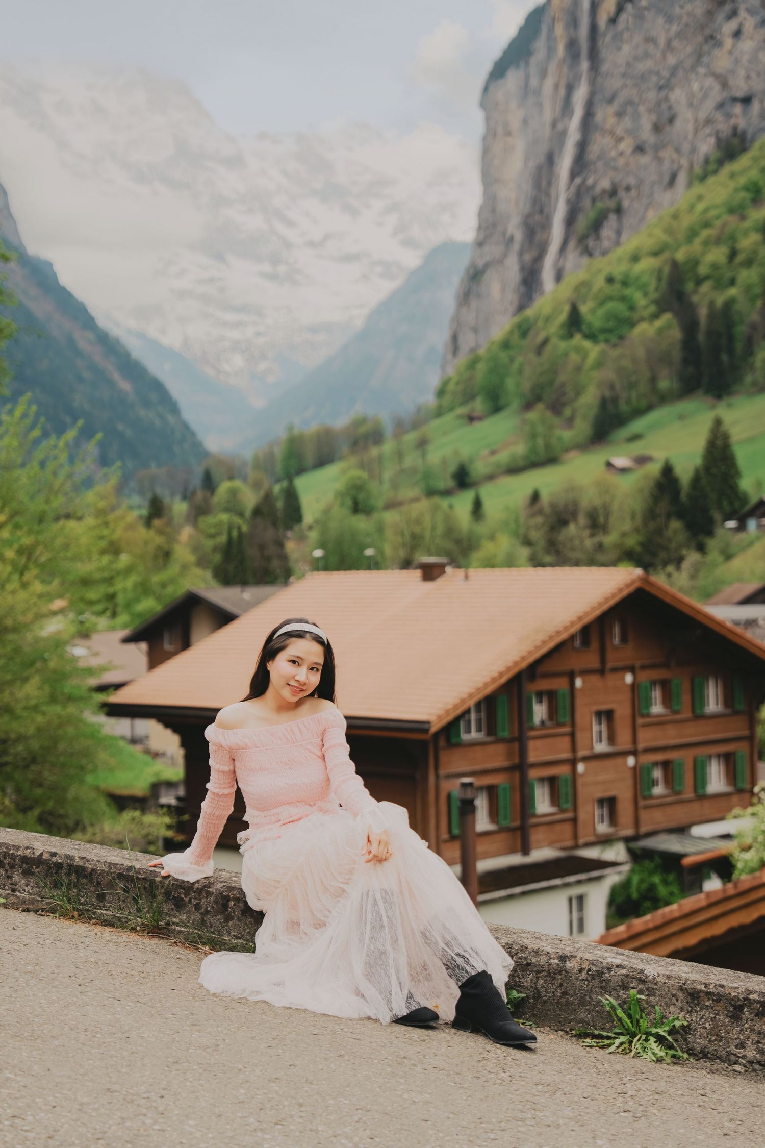 Woman in pink dress sitting on stone wall with Swiss Alps, waterfall, and Lauterbrunnen valley behind her.