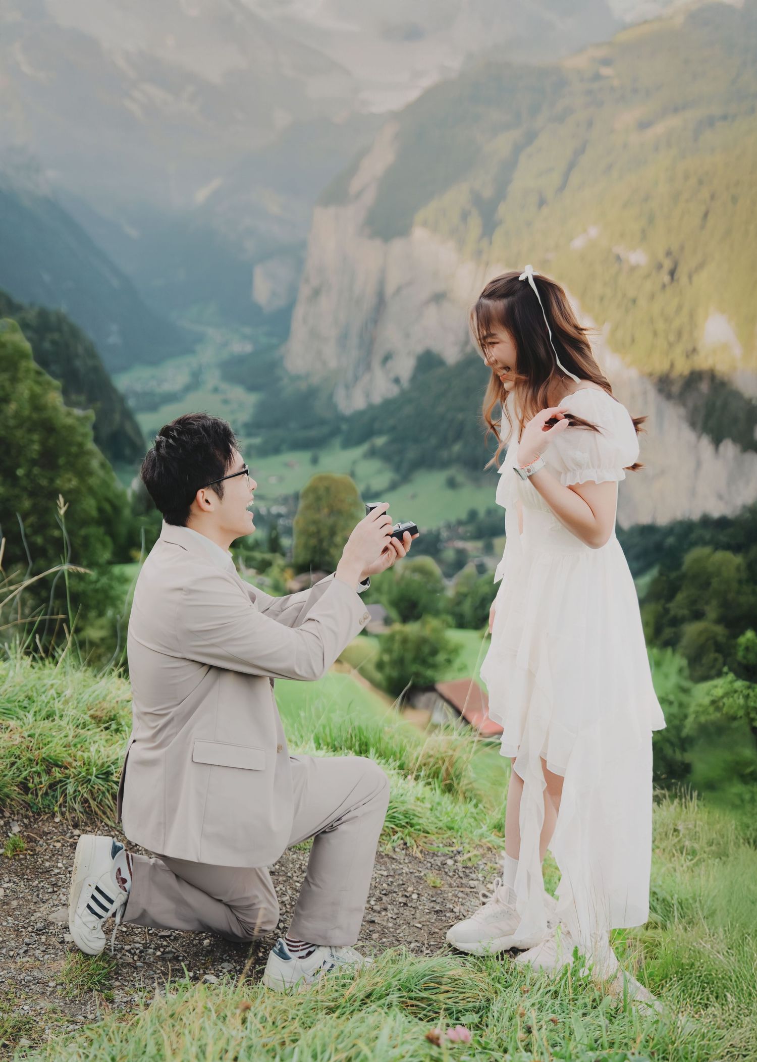 Man proposing on one knee to woman in white dress with stunning Swiss valley landscape behind them.