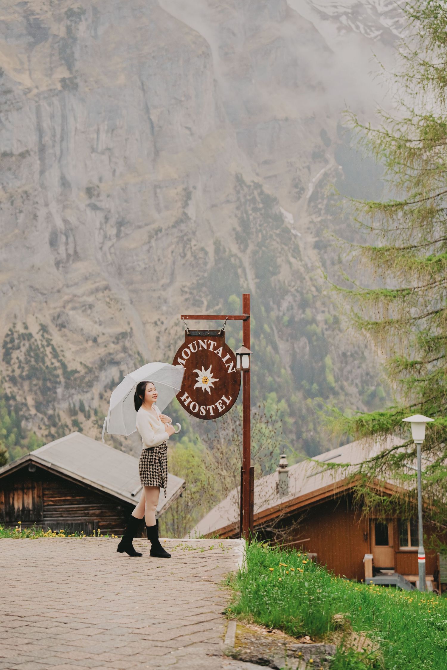 Woman with white umbrella standing beside Mountain Hostel sign in misty alpine village.