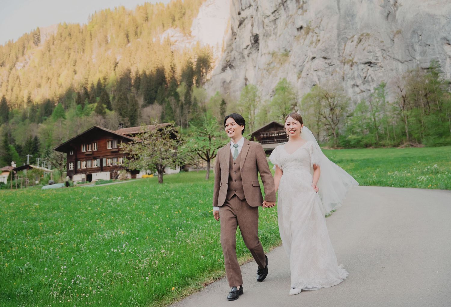 Bride and groom walk hand in hand along a path with stunning Swiss rocky cliffs and chalets behind them.