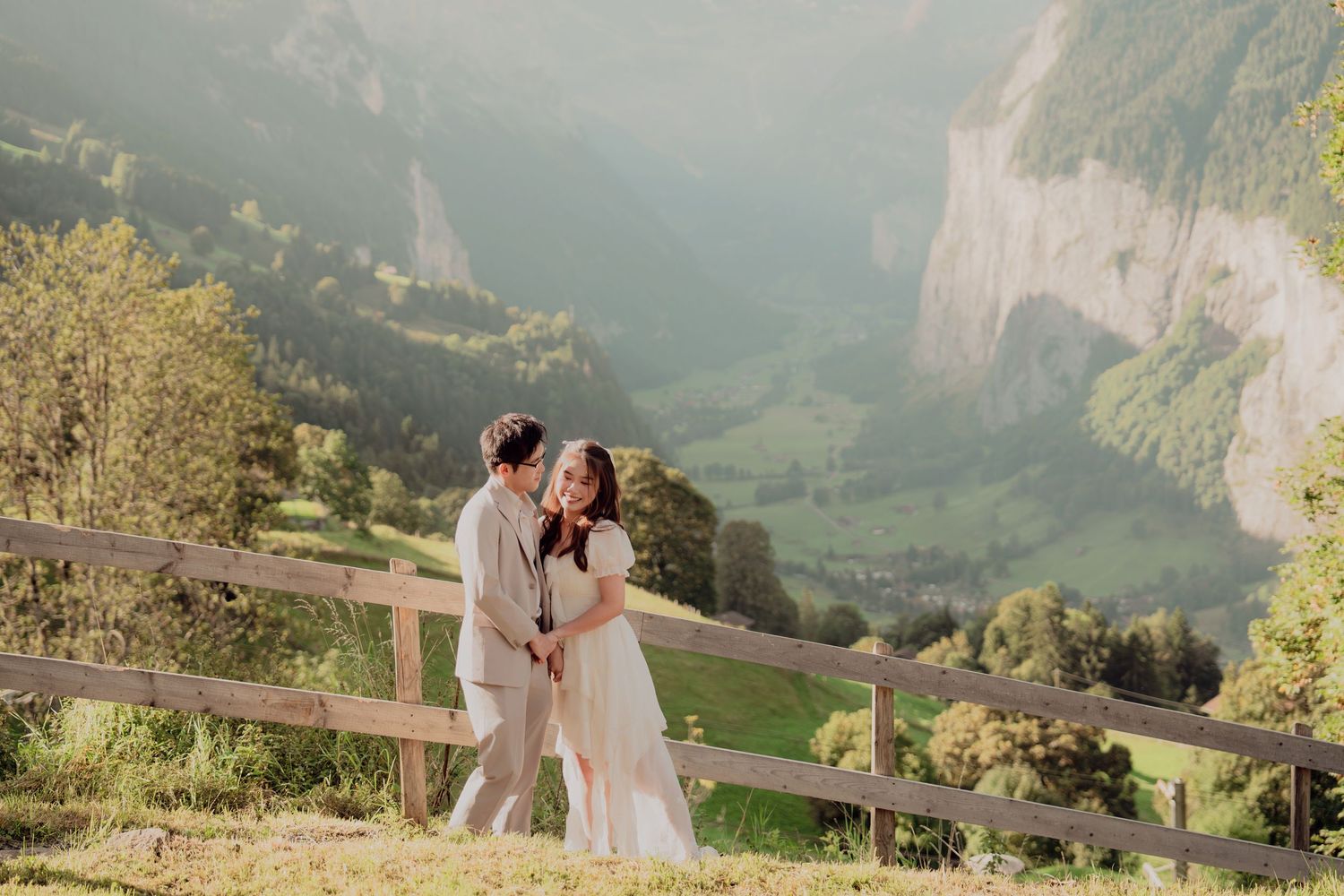Couple sharing a romantic moment by a wooden fence with lush Swiss alpine valley scenery.
