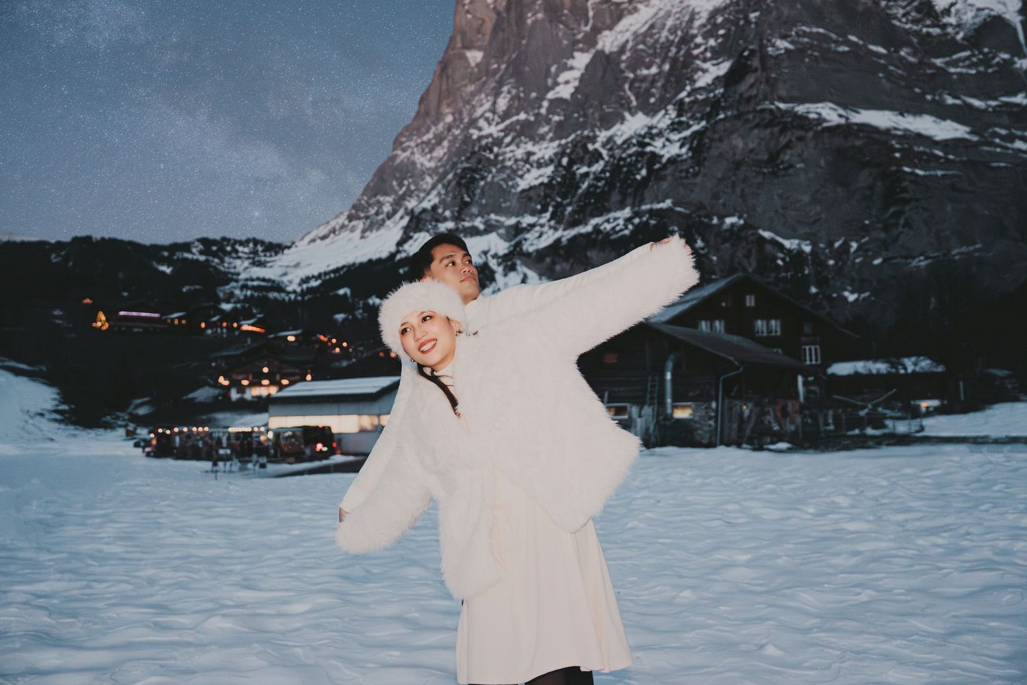 Woman in white fur coat being lifted by partner in snowy alpine setting under night sky.
