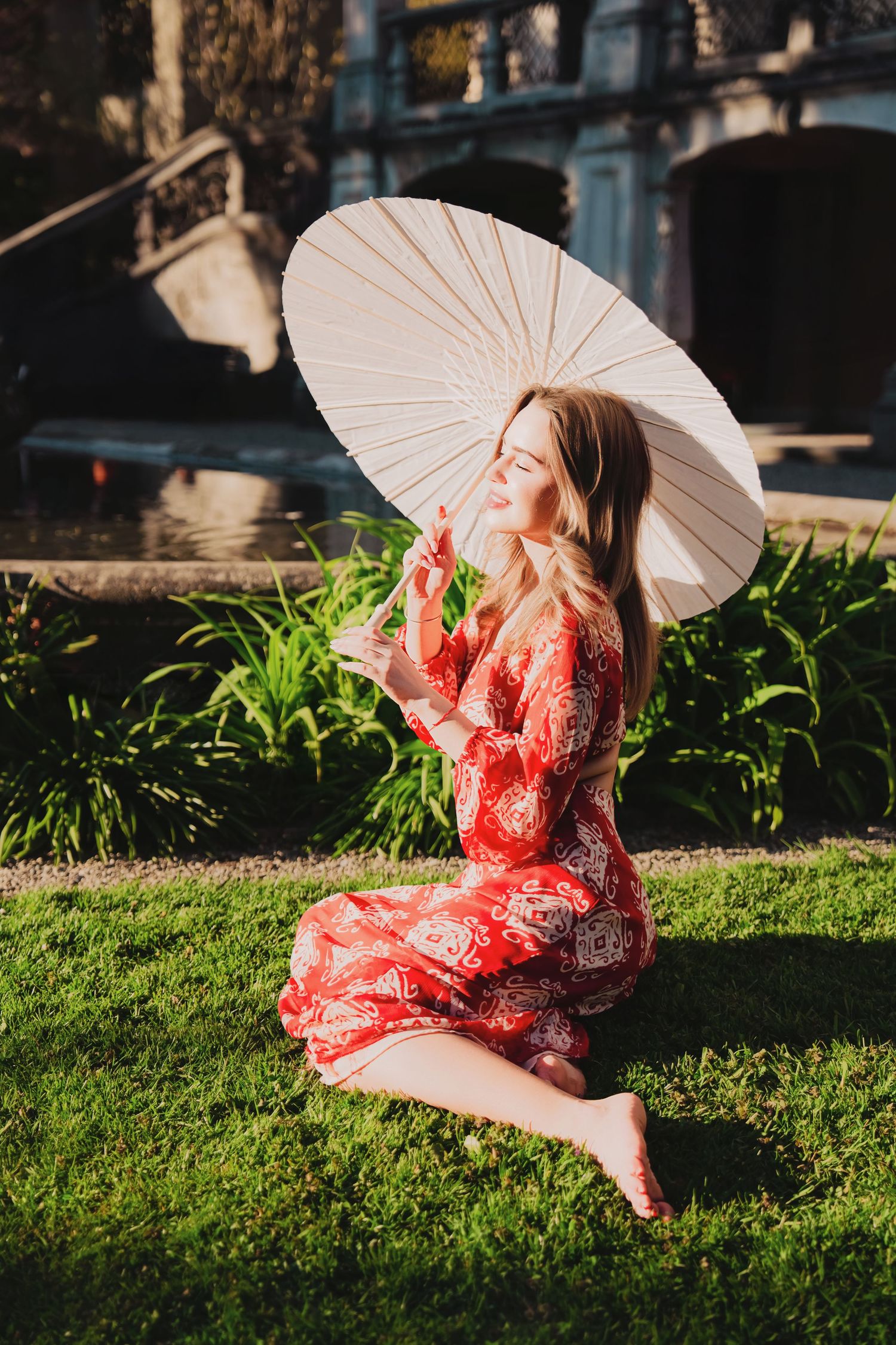 Woman in red floral dress sits barefoot on grass holding a white paper parasol umbrella outdoors.