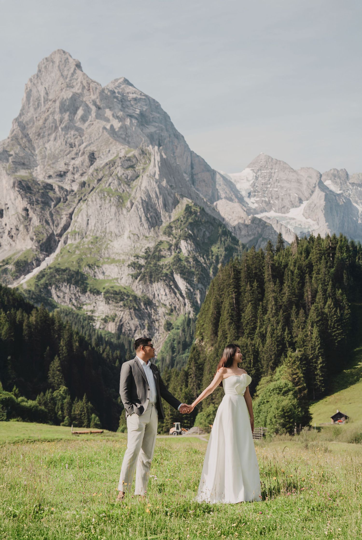 Wedding couple holds hands in green alpine meadow with dramatic rocky Swiss mountain peaks behind.