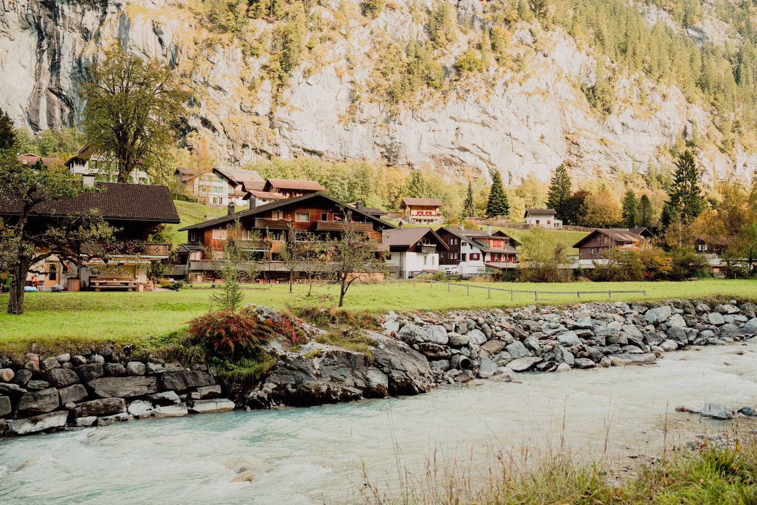 Swiss alpine village with traditional wooden chalets beside a turquoise glacial river with rocky banks and mountain backdrop.