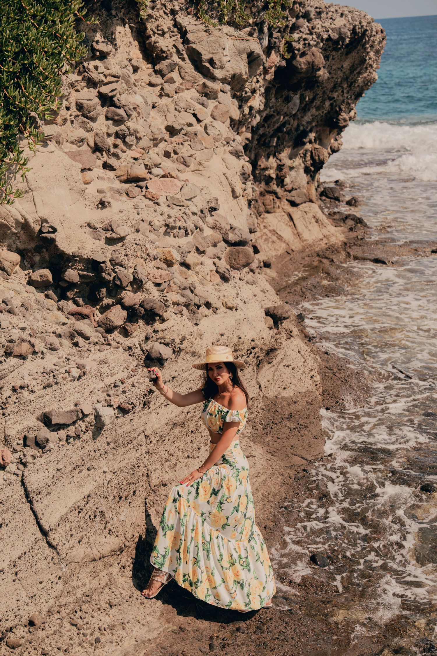 Woman in floral dress and straw hat posing on sandy beach beside rocky cliffs.