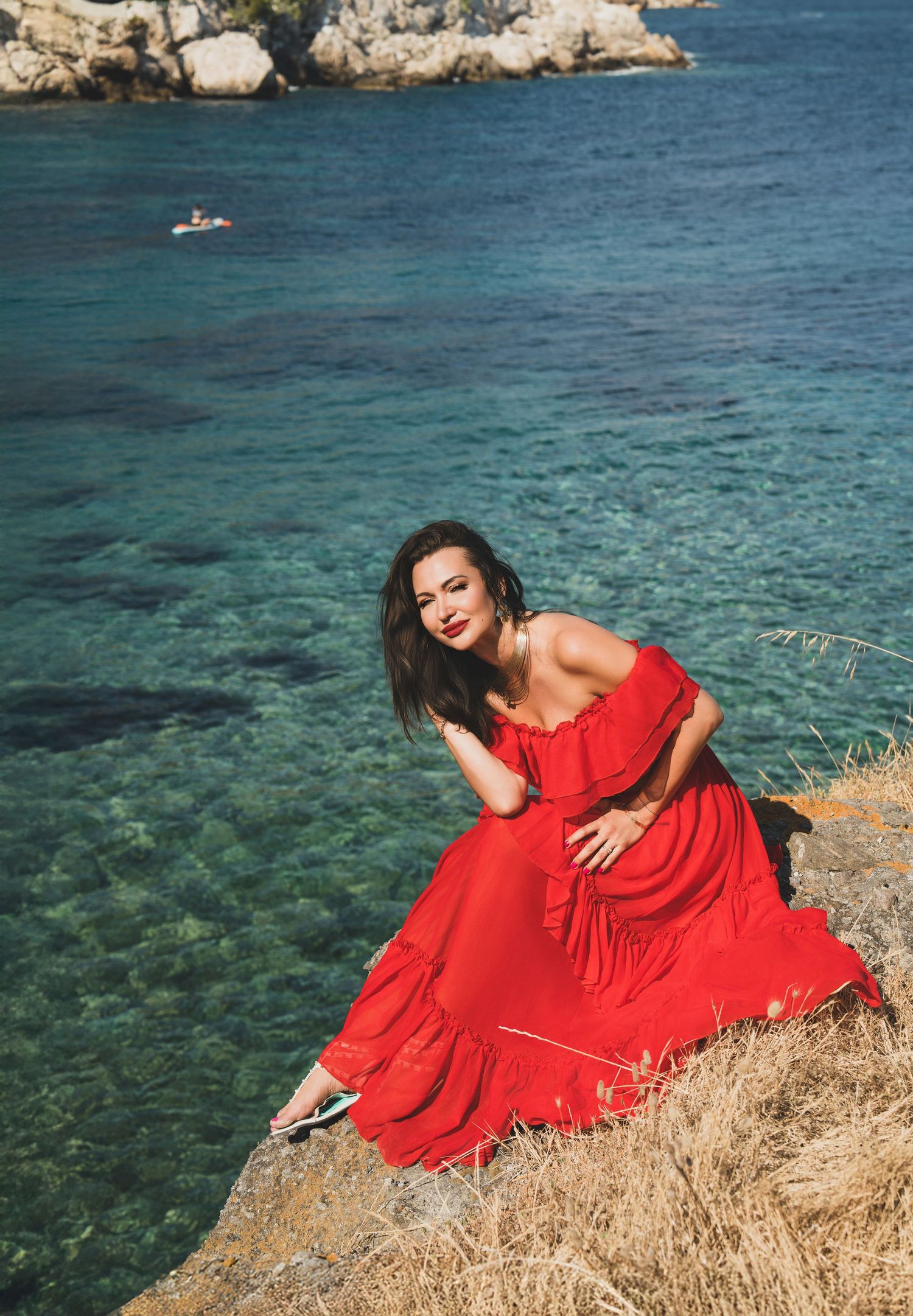 Woman in red off-shoulder dress sitting on rocky cliff by turquoise Mediterranean sea.