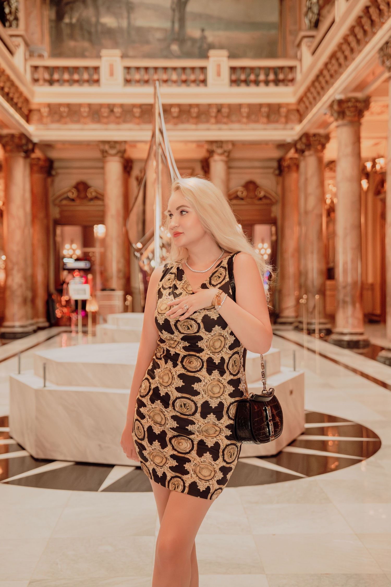 Elegant blonde woman in black and gold patterned dress posing inside a luxurious ornate marble interior.