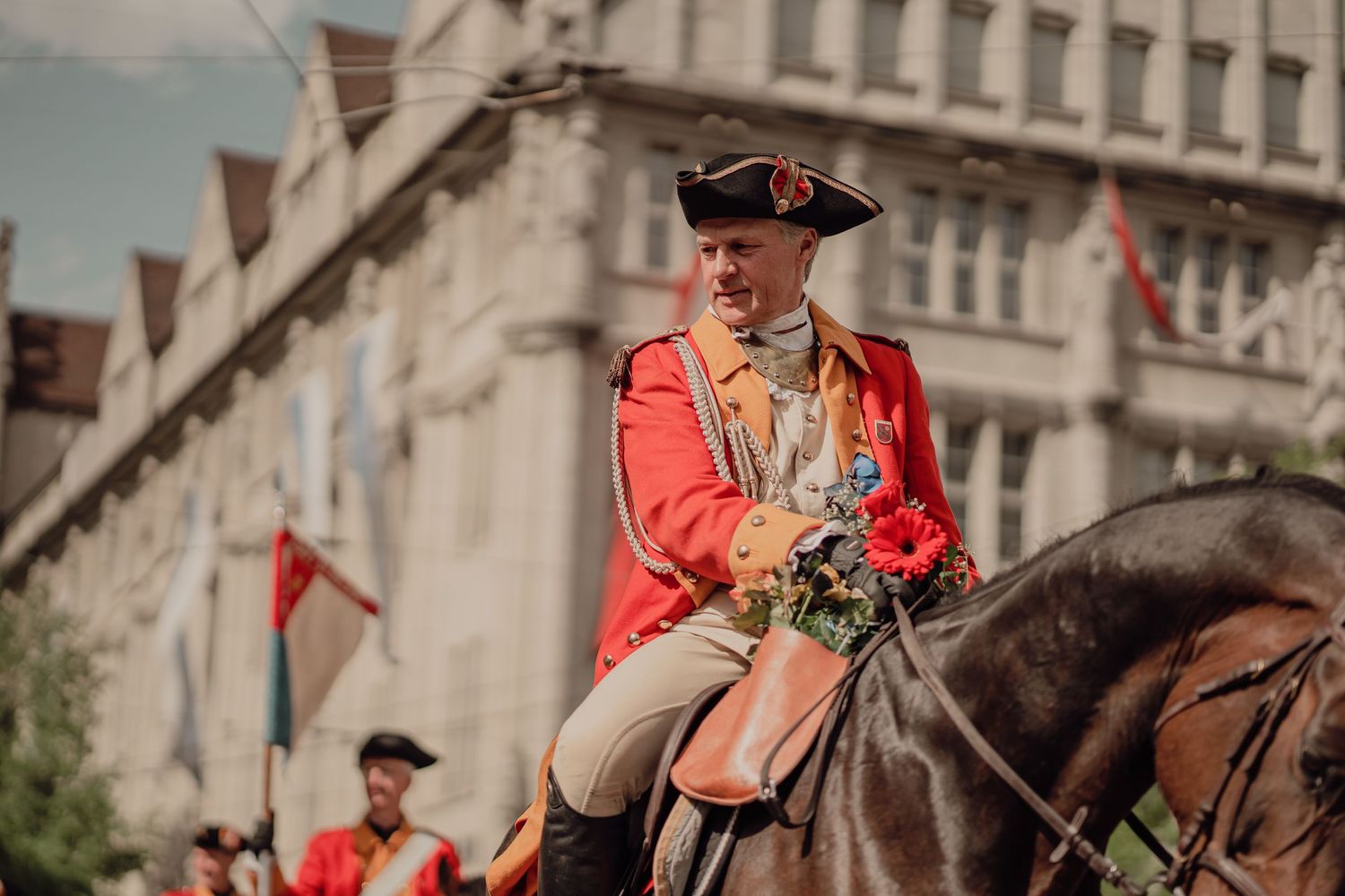Man in red 18th-century military uniform rides a horse holding flowers in a historical parade.