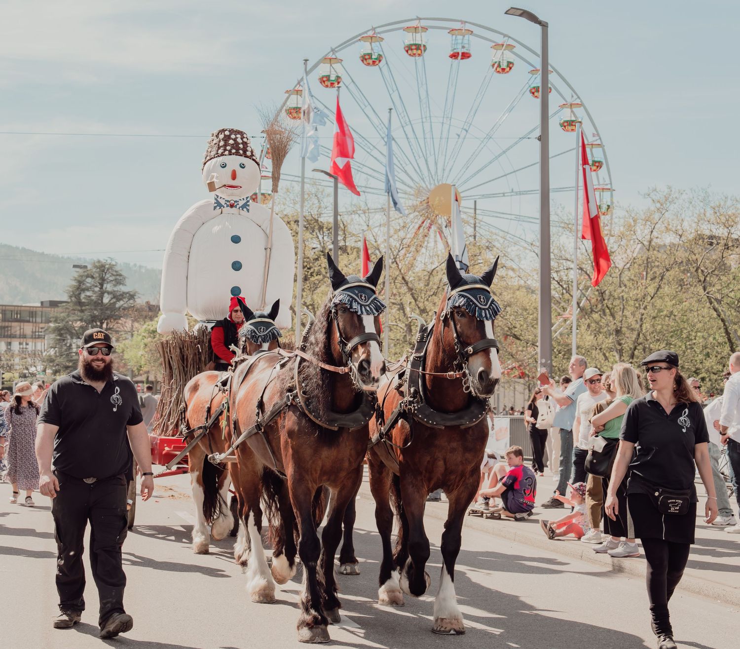 Draft horses pulling a snowman float past a Ferris wheel at a Swiss spring festival.