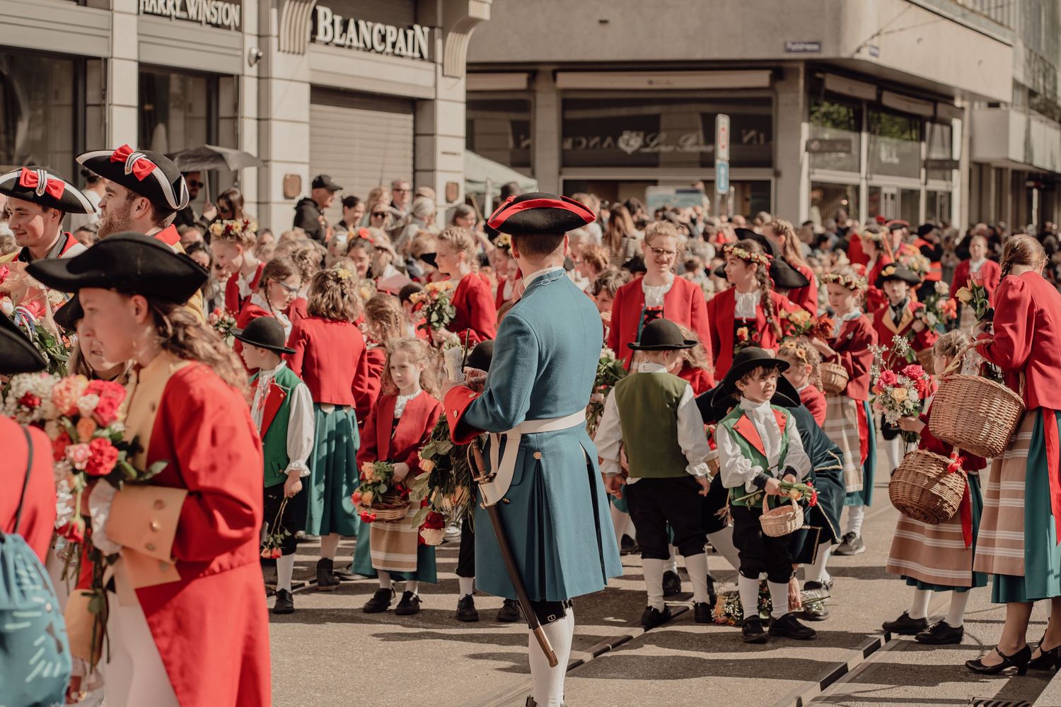Participants in red and teal traditional folk costumes gather during a colorful outdoor parade event.