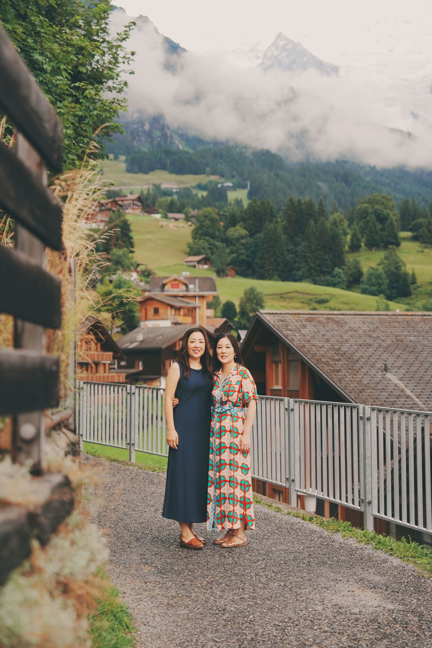Two Asian women pose together on a gravel path with misty Swiss Alps mountains behind them.