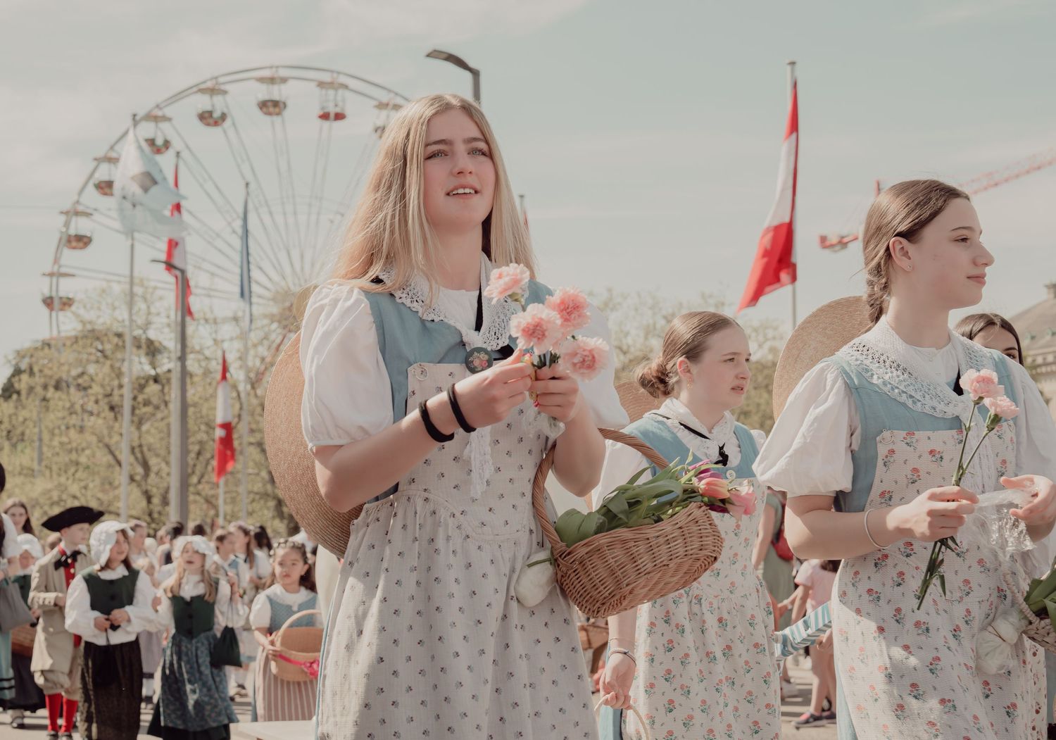 Young women in traditional folk costumes carry flower baskets in a sunny outdoor parade.