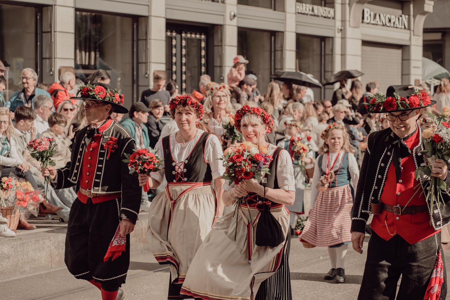 Group of performers in traditional black and red Polish folk costumes carrying red flower bouquets in a street parade.