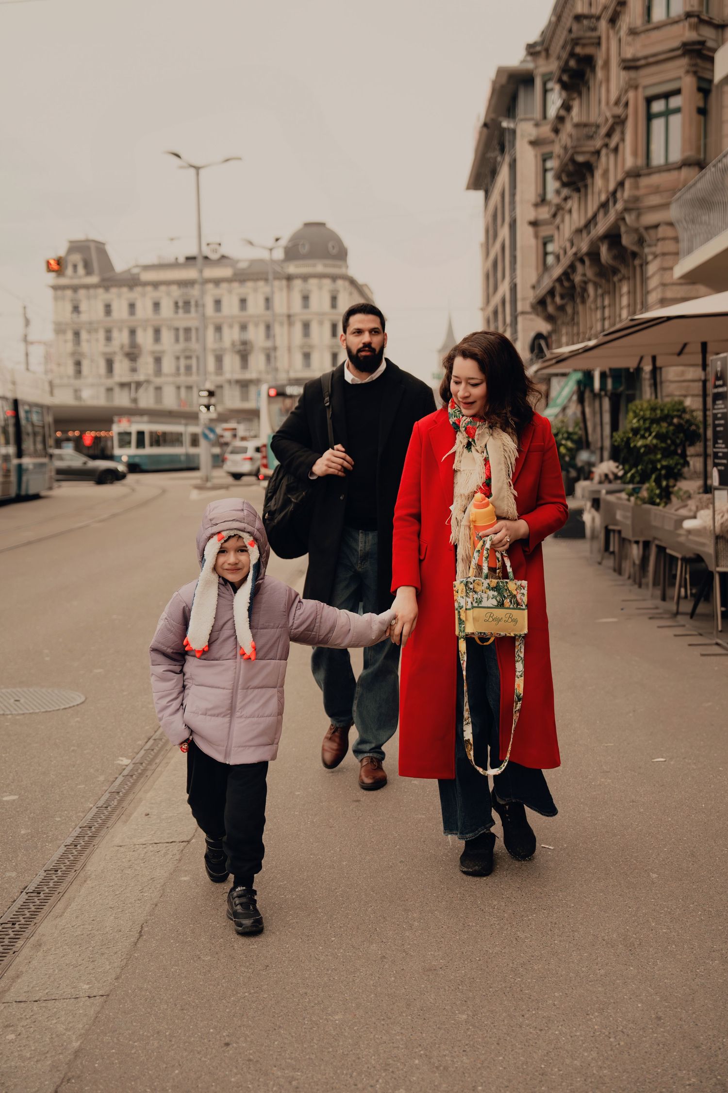 Family strolling on a city sidewalk, young child in pink jacket holding mother's hand in red coat.