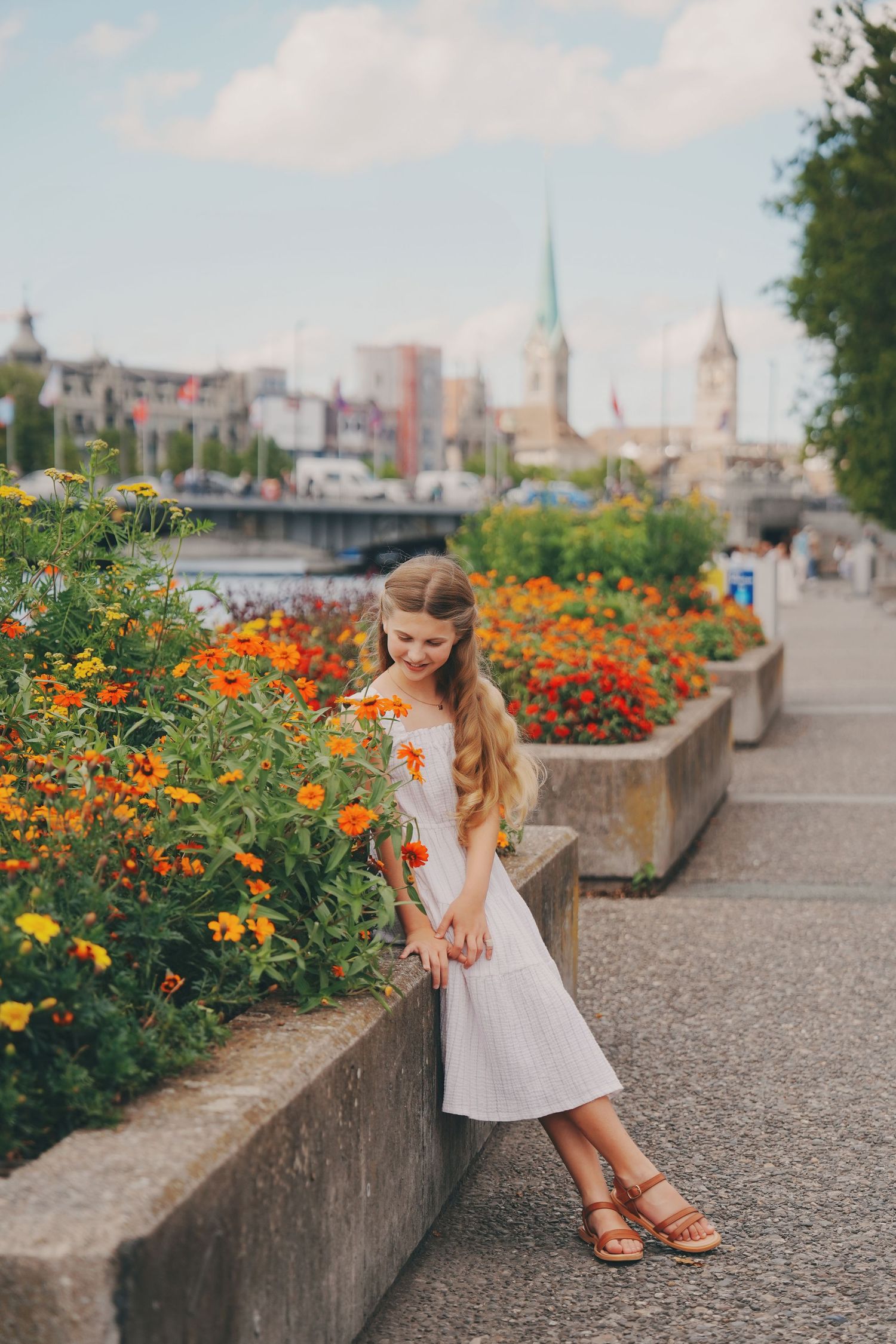 Young girl in white dress standing by colorful orange flowers along a scenic Zurich waterfront promenade.