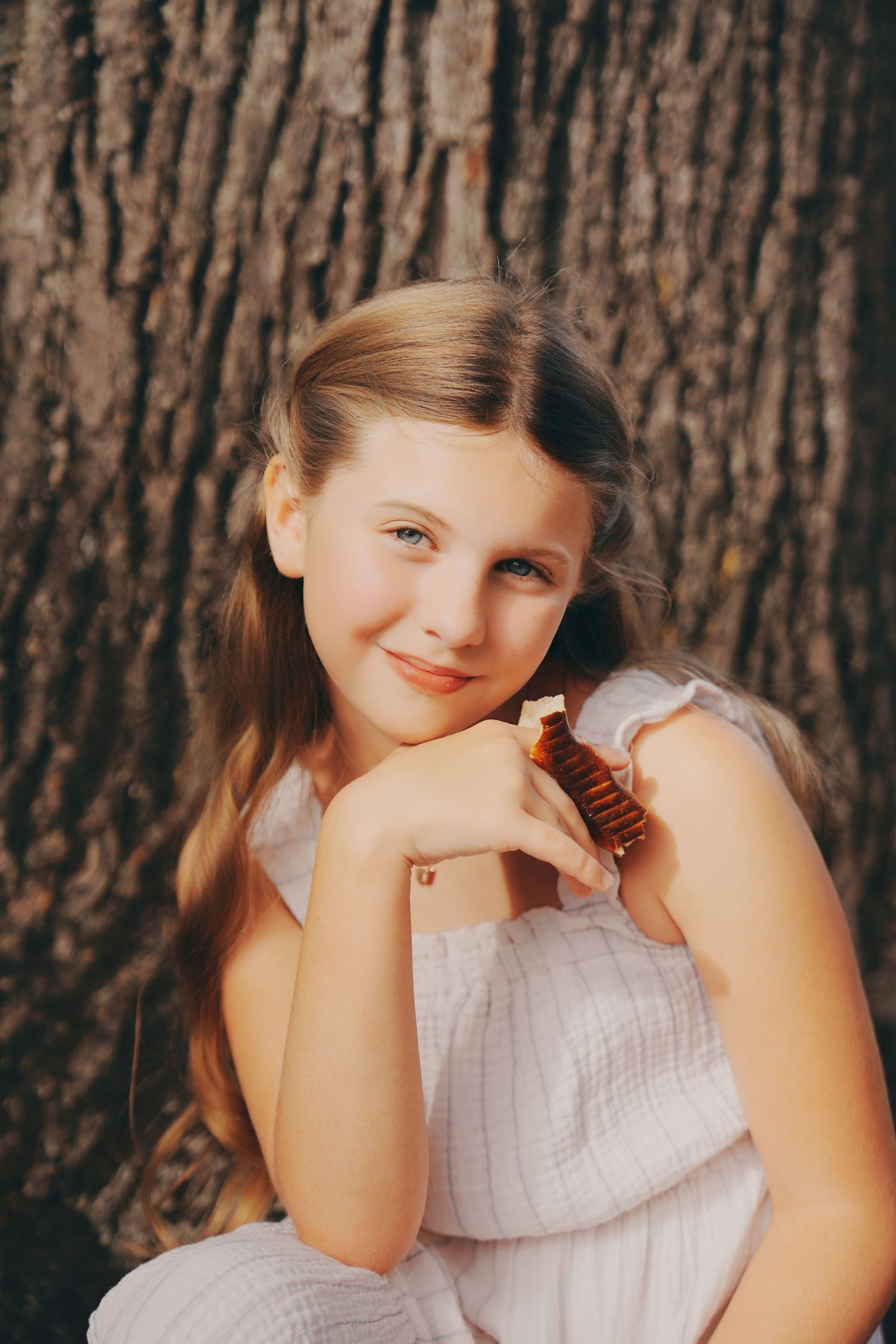 Smiling young girl in white dress leaning against textured tree bark outdoors.