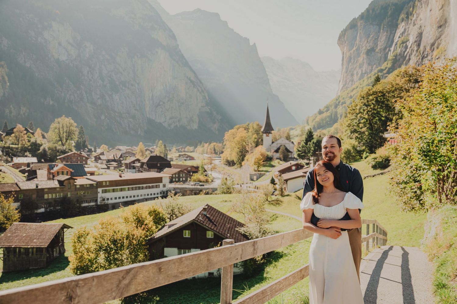 A couple embracing on a balcony with a breathtaking Swiss alpine village and misty mountains in background.