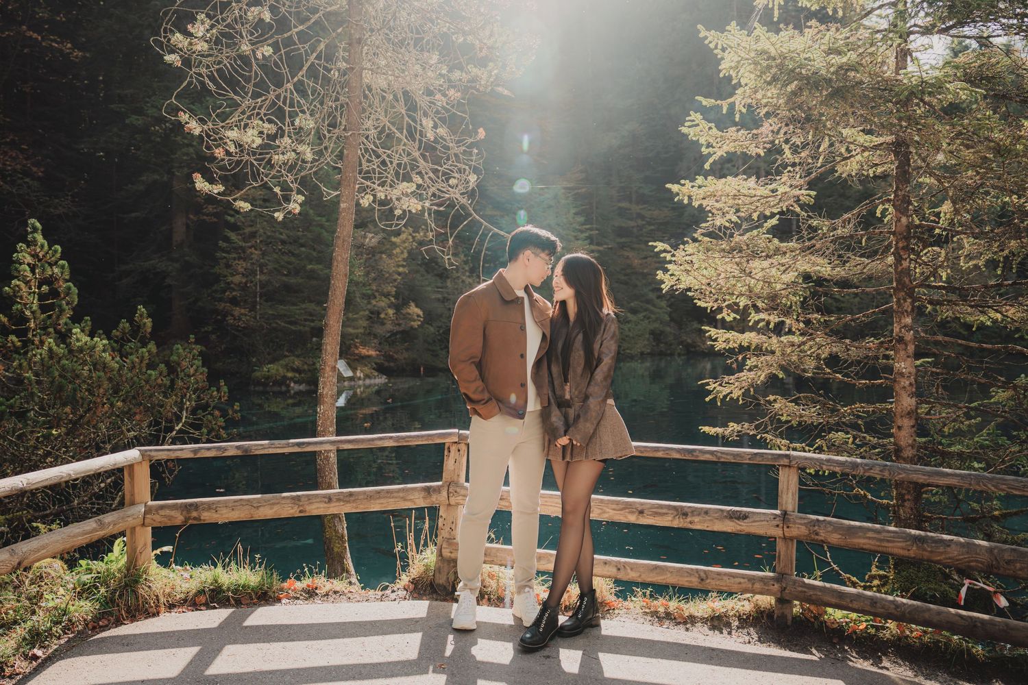 Couple sharing a romantic moment on a wooden bridge surrounded by tall pine trees and a serene blue lake.