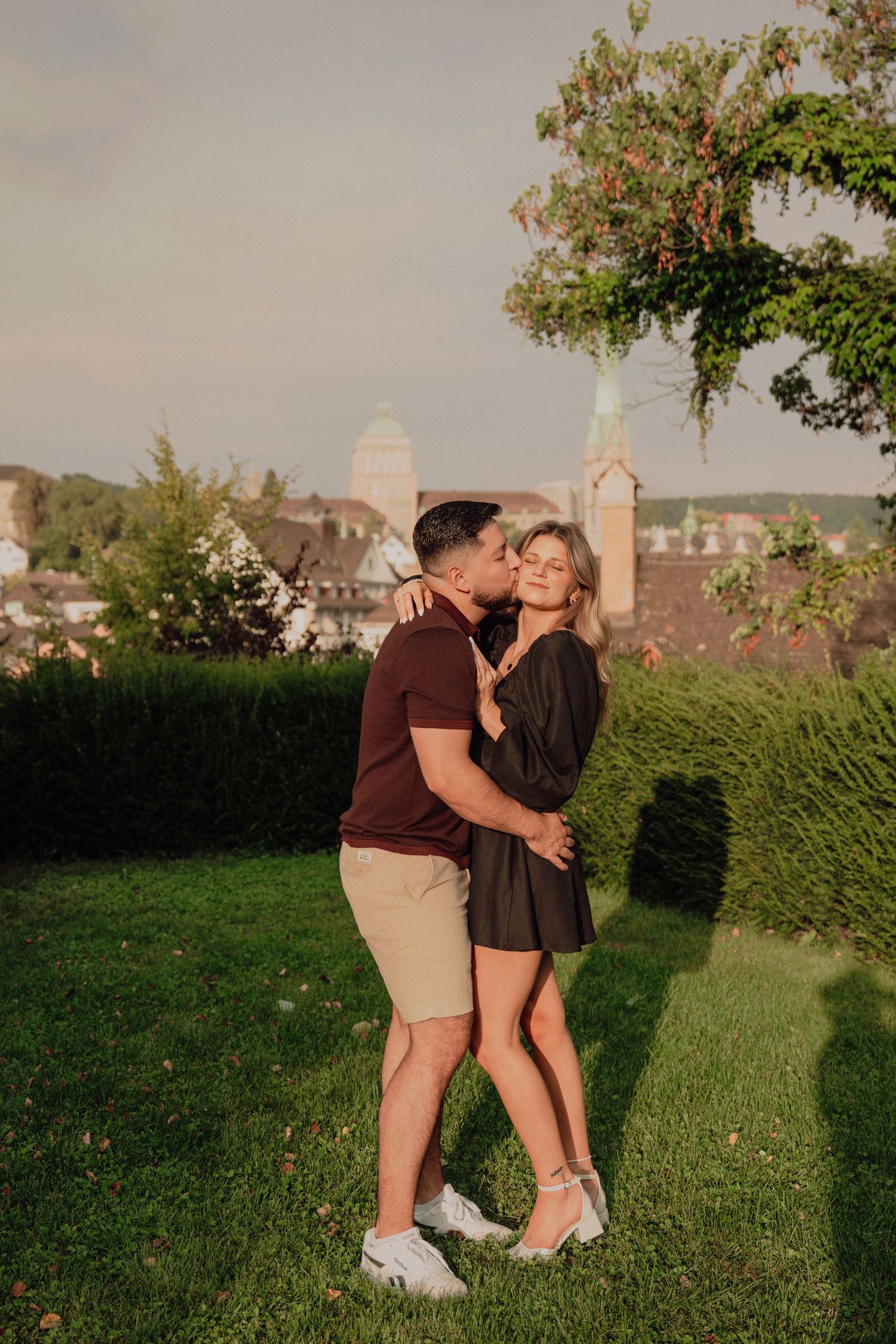 Couple sharing a romantic kiss on a green lawn with Zurich's historic church towers visible in background.