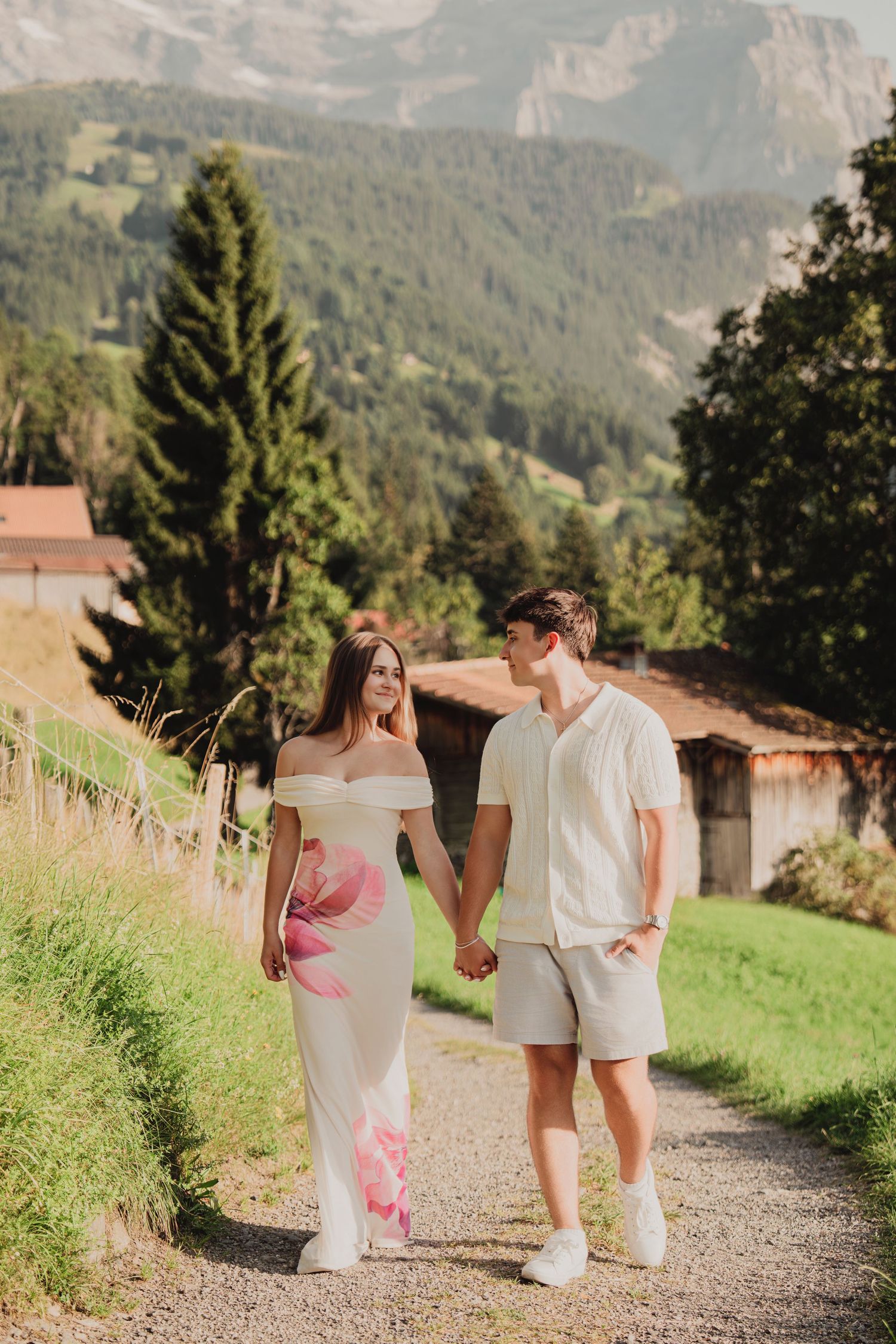 A couple holds hands walking on a gravel path through a scenic alpine village setting.