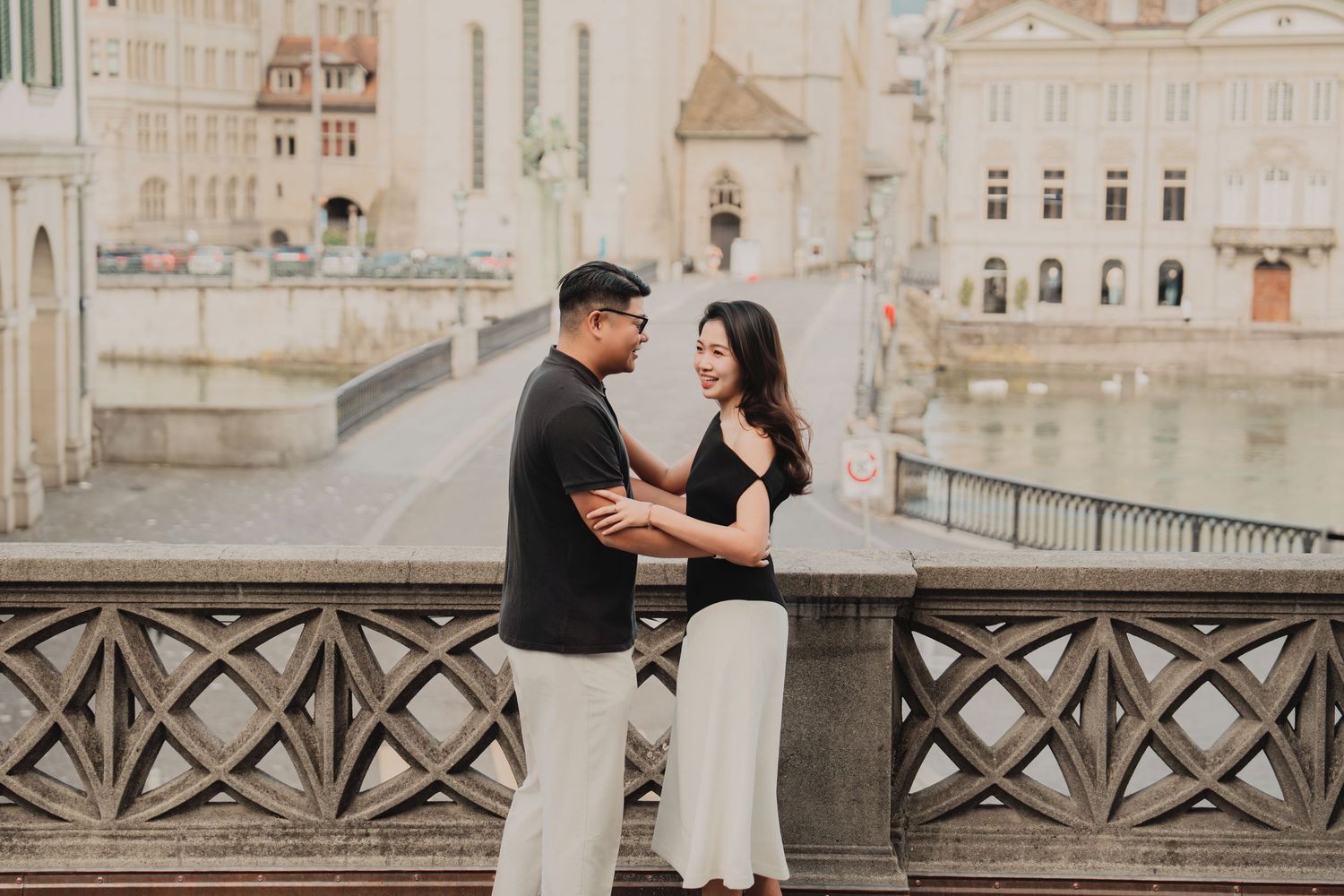 Couple embracing and smiling at each other on a Zurich bridge with old town architecture behind.