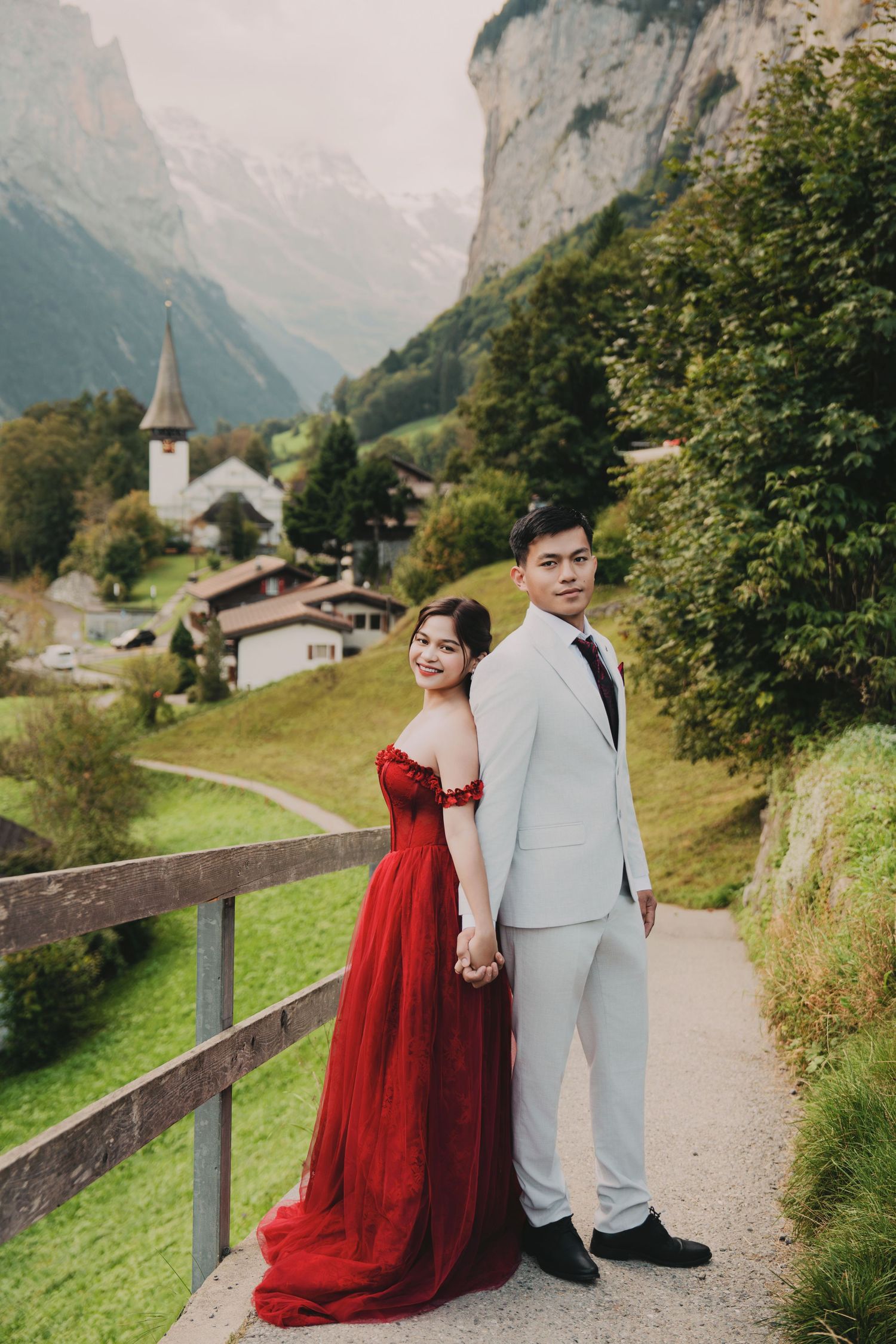 Couple posing hand-in-hand on Swiss village path with dramatic mountain cliffs and church tower.