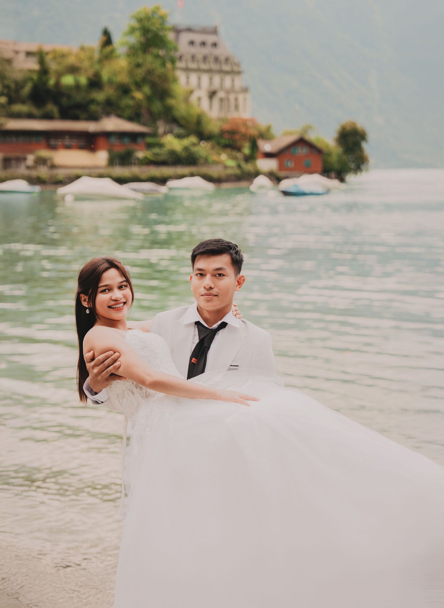 Elegant couple in wedding attire posing by a calm alpine lake with a grand historic building behind.