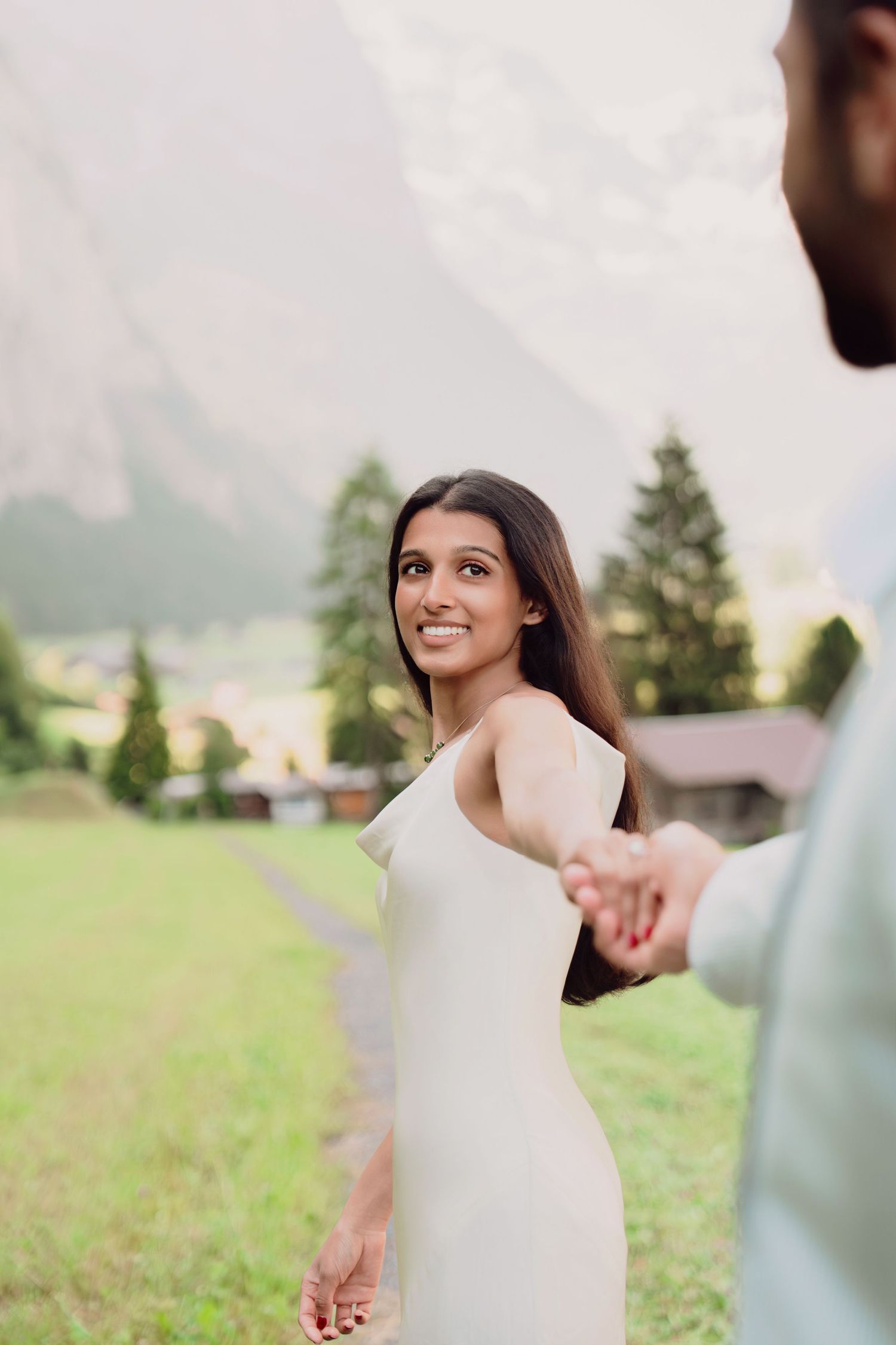 Smiling bride in white slip dress holding hands with groom in lush Alpine meadow setting.