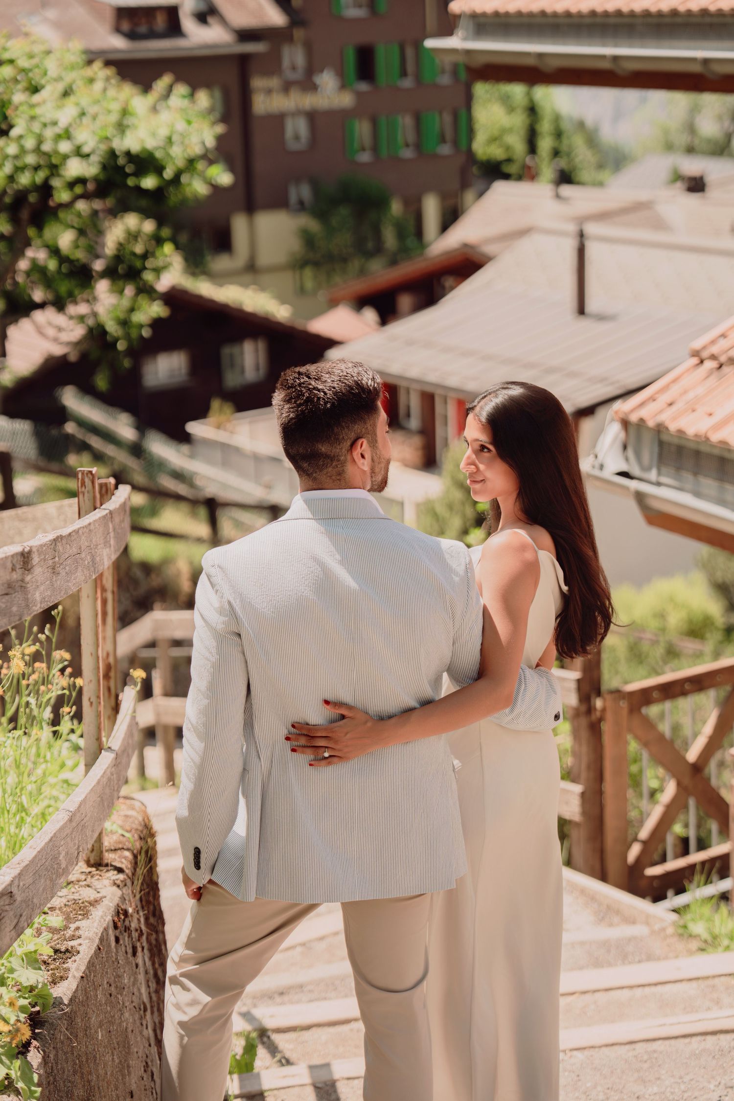 Couple sharing a tender moment on rustic wooden stairs overlooking charming Swiss village buildings on a sunny day.