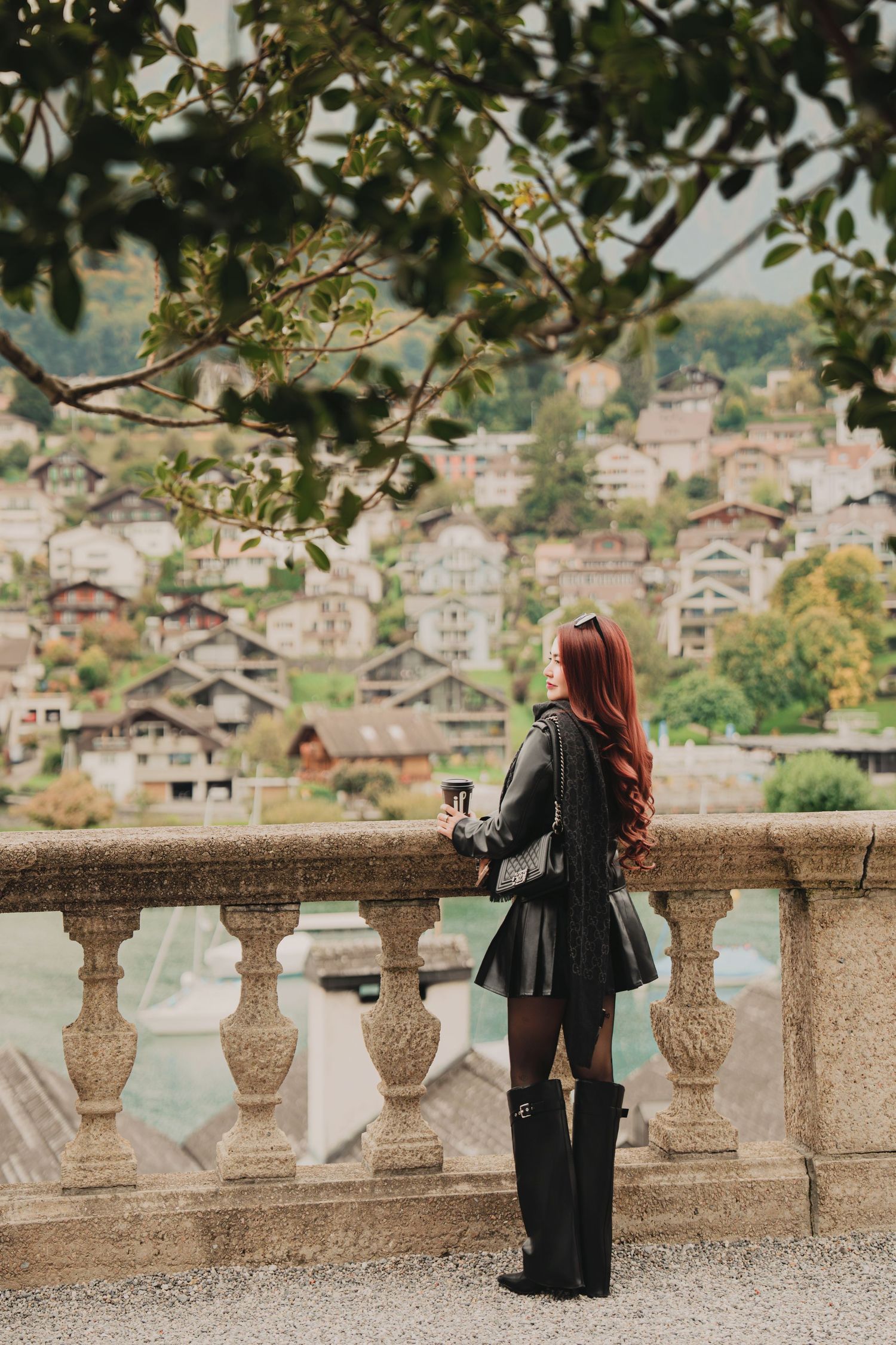 Stylish red-haired woman in black leather mini skirt and tall boots holds coffee on ornate stone balcony overlooking Swiss town.