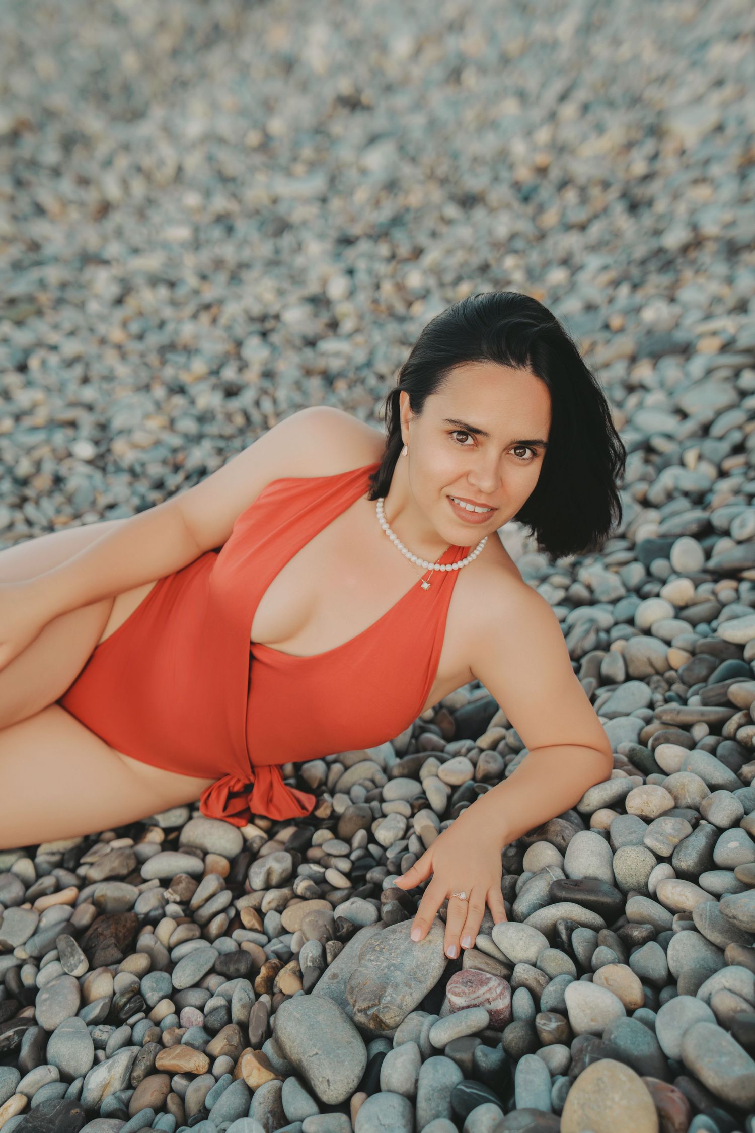Woman in red swimsuit posing on pebble beach, smiling at camera.