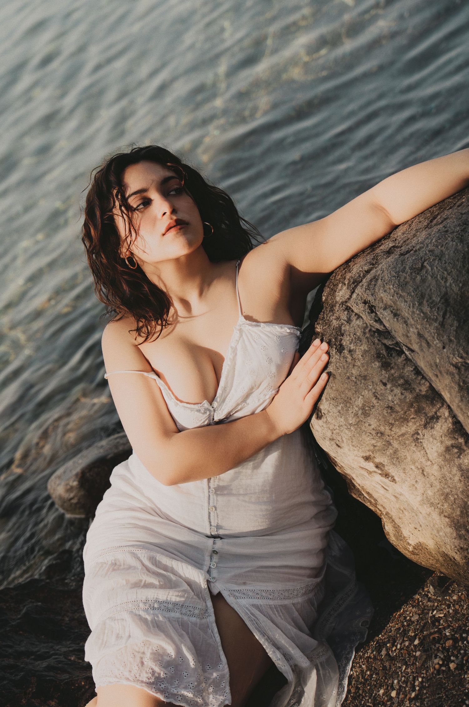 Woman in wet white dress leaning against a rock by the sea.