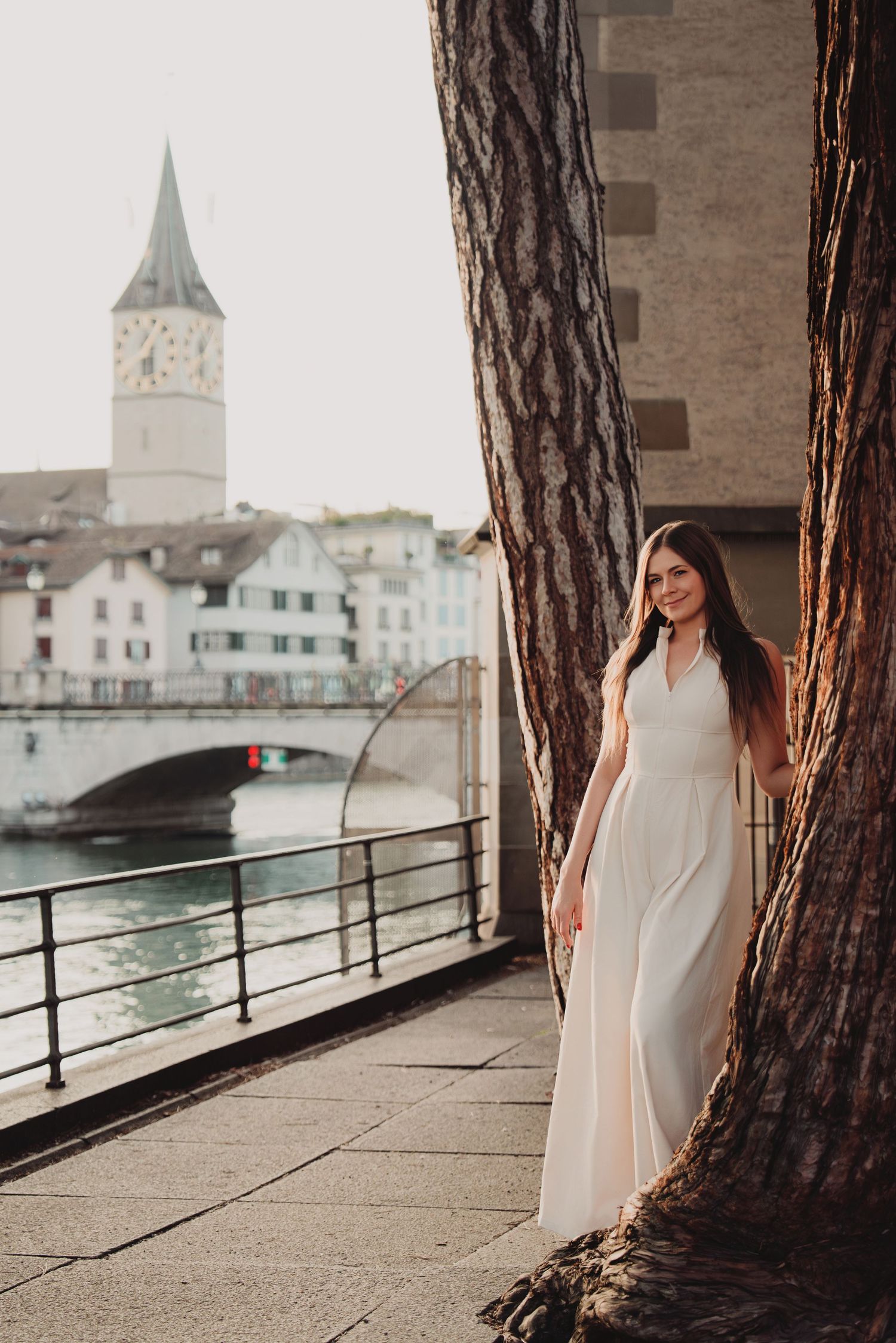 Smiling woman in white dress posing between large trees along Zurich riverside with church tower view.
