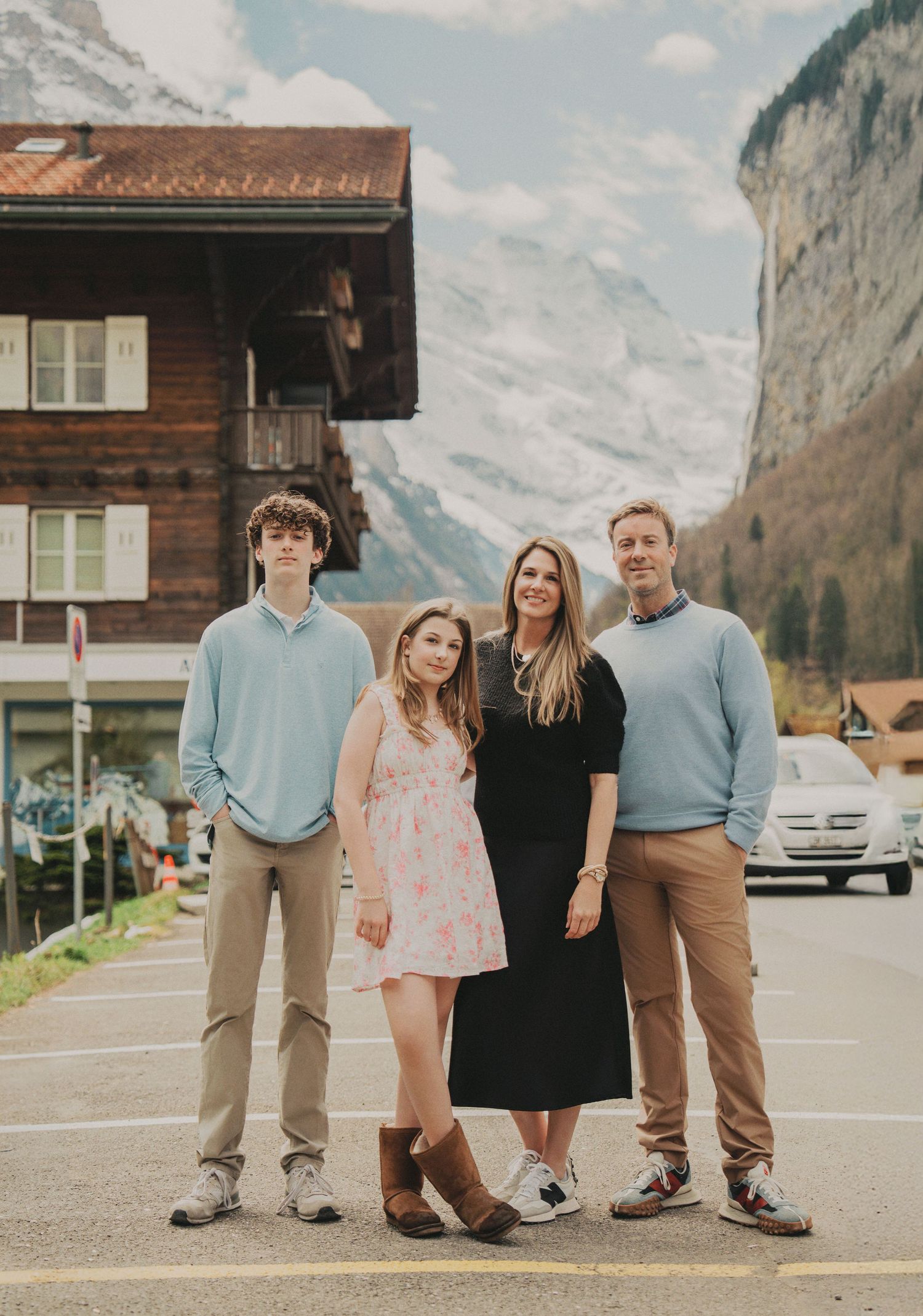 Family of four posing on a street in Swiss Alps village with snow-capped mountains behind.