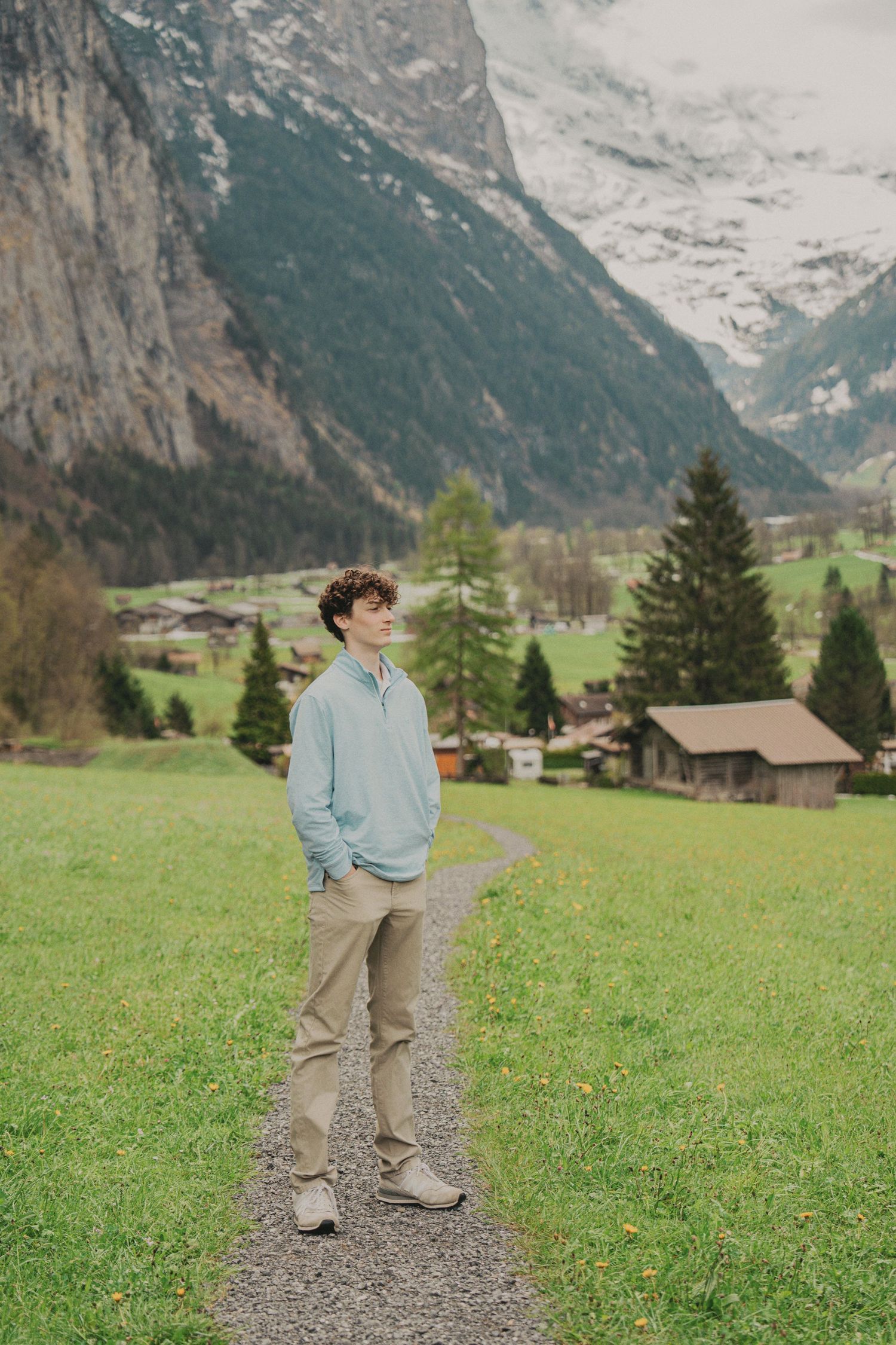 Teen boy in light blue sweater standing on mountain path gazing at snow-capped Swiss Alps.