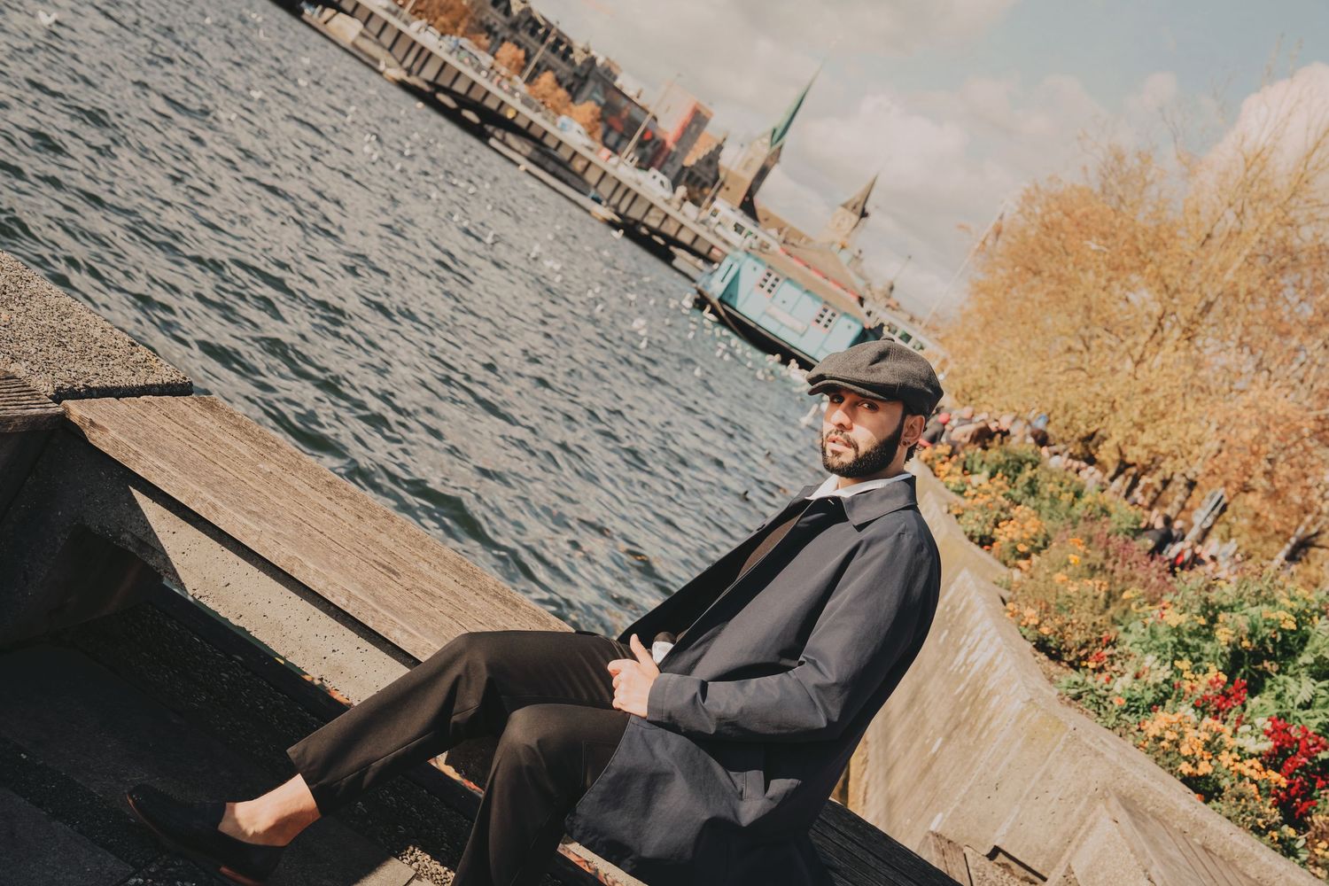 Bearded man in gray blazer and flat cap relaxes on wooden bench by scenic autumn waterfront.