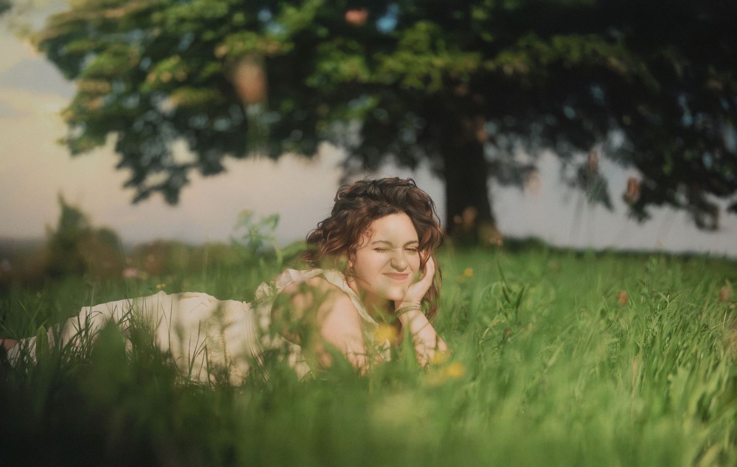 Smiling woman lying in green grass field beneath a large tree in warm golden light.