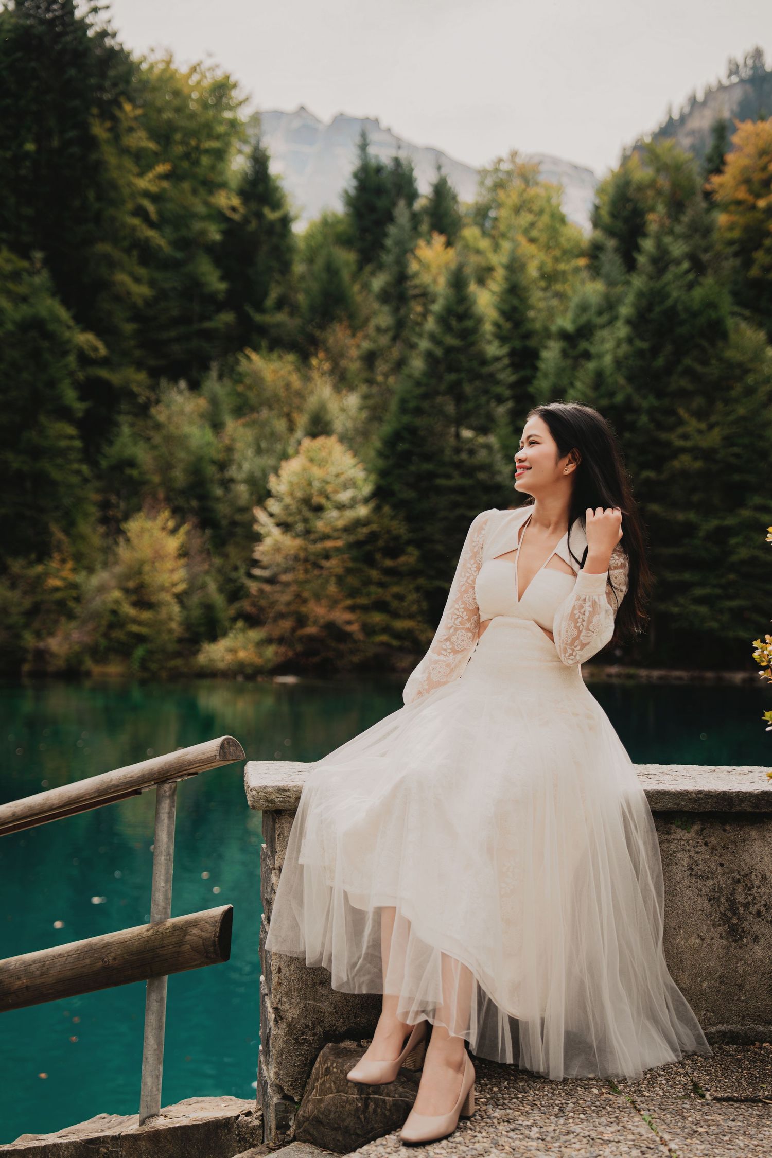 Smiling woman in white tulle dress sits on stone railing by turquoise mountain lake.