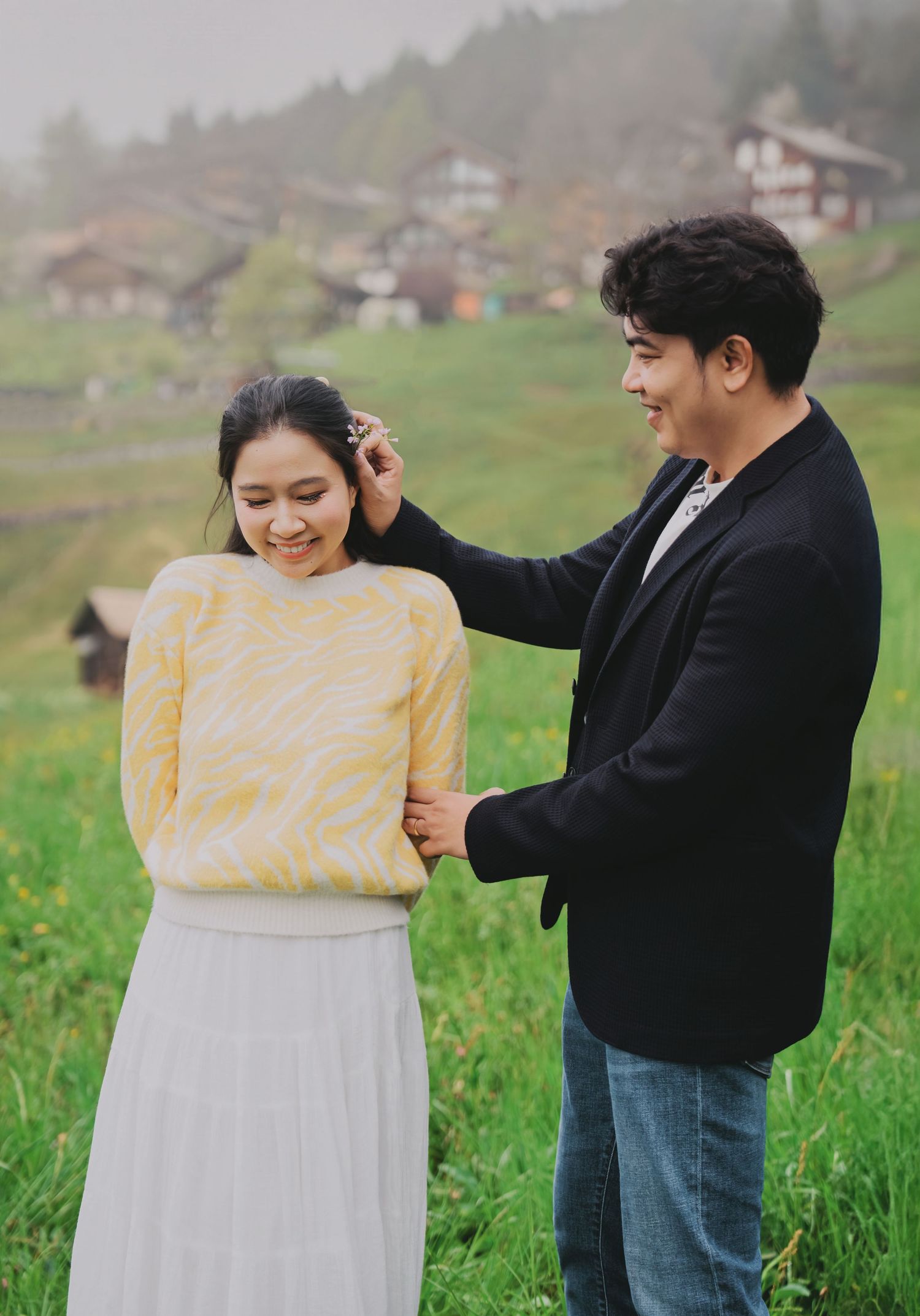 Man gently fixing womans hair while she smiles on a green meadow with misty village backdrop.