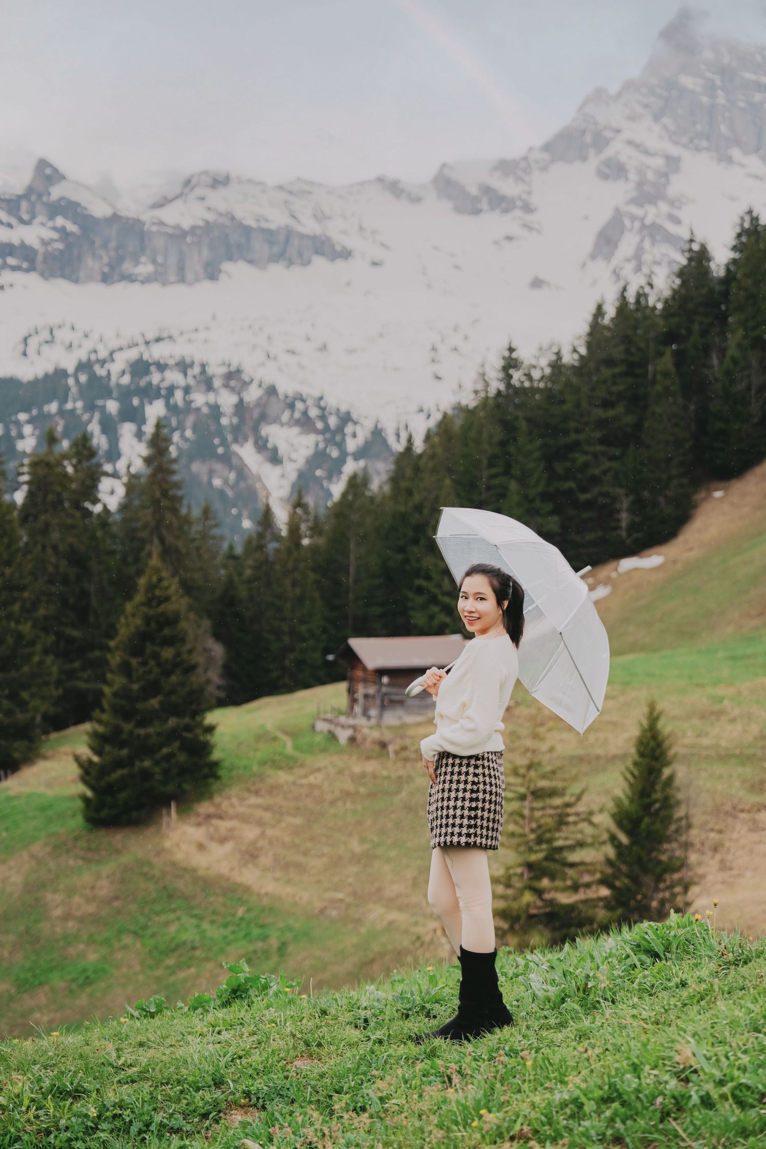 Smiling woman holds clear umbrella on alpine meadow with snow-capped mountains and pine forest backdrop.