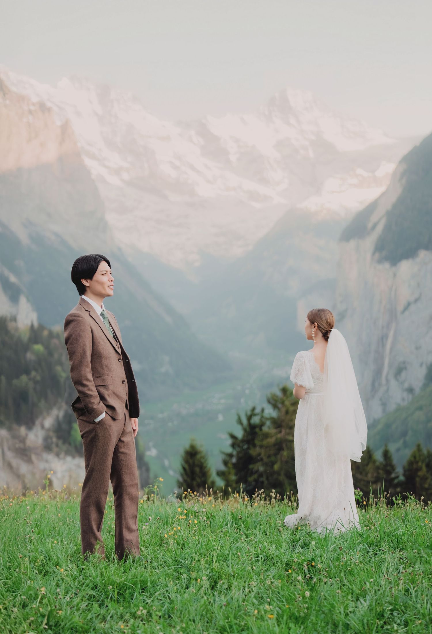 Bride and groom standing apart on green hillside with dramatic Swiss Alps valley view.