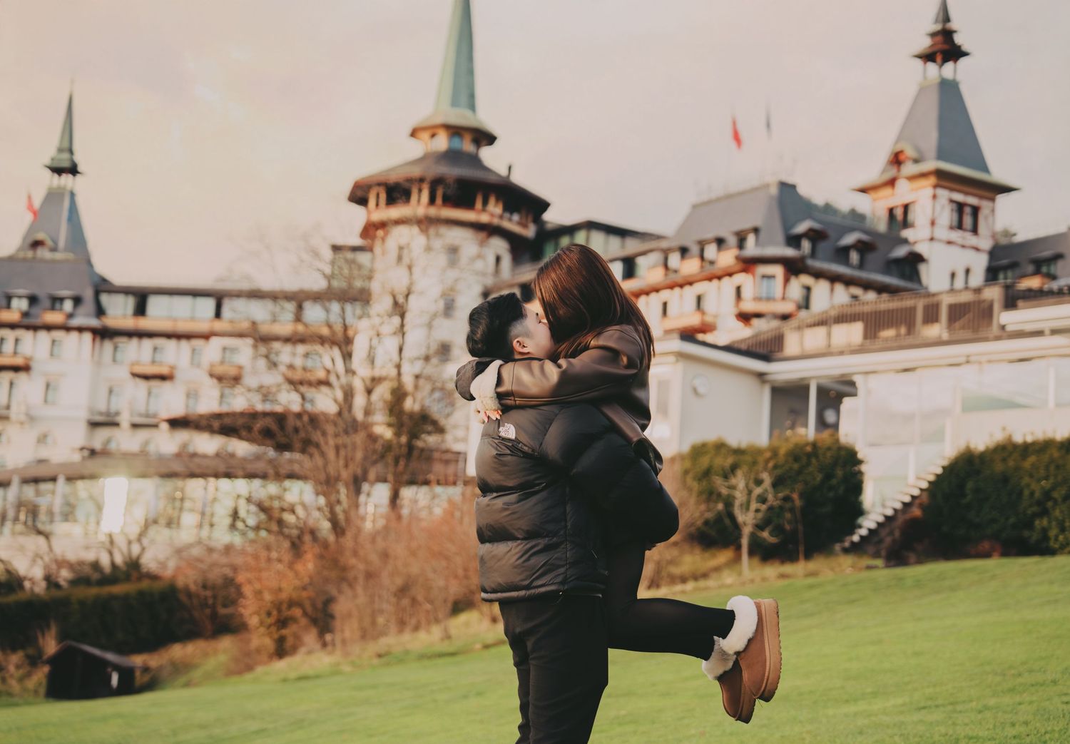 Man joyfully lifting woman on green lawn with grand European château in background.