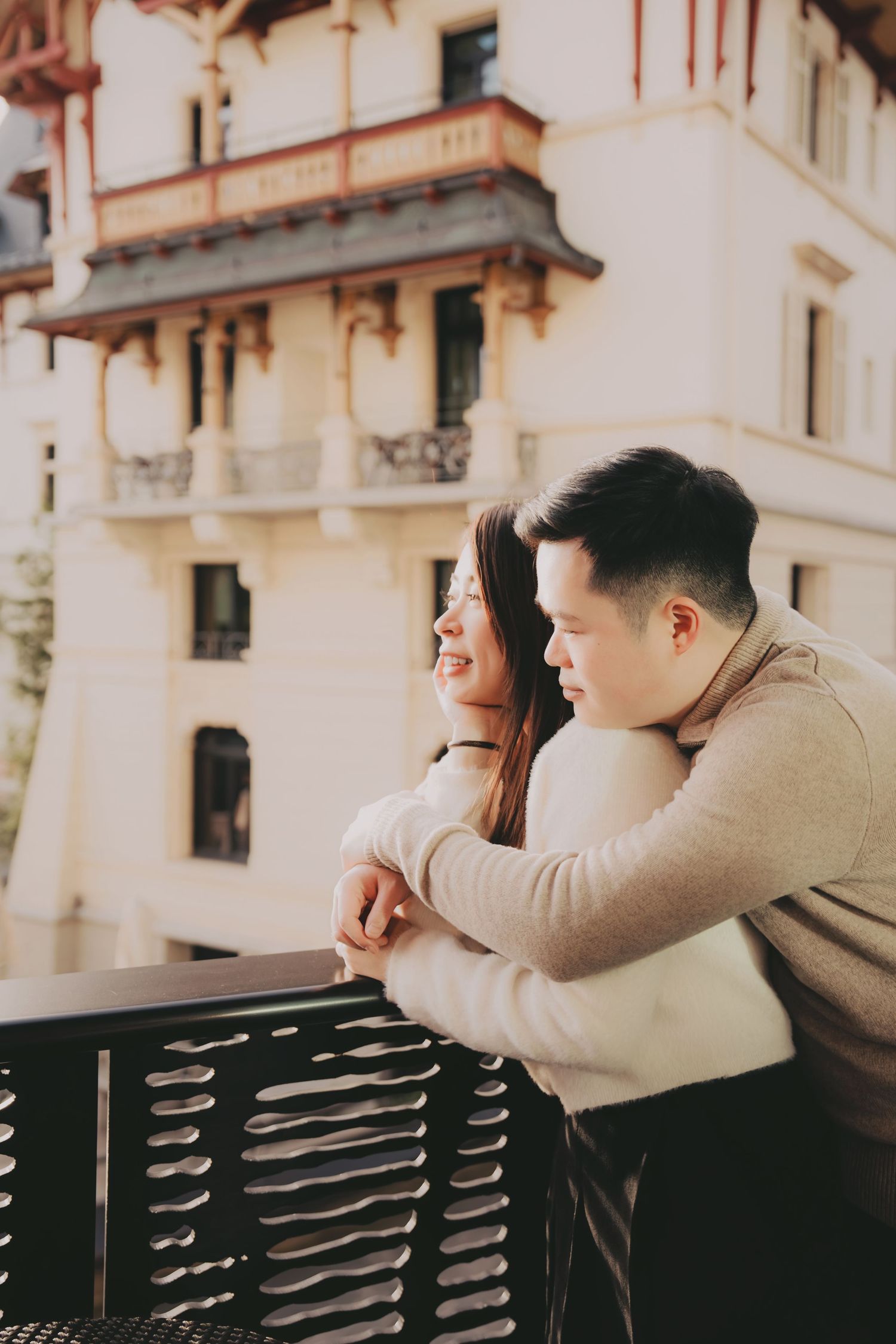 Happy Asian couple leaning on a balcony railing smiling with an ornate historic building behind them.