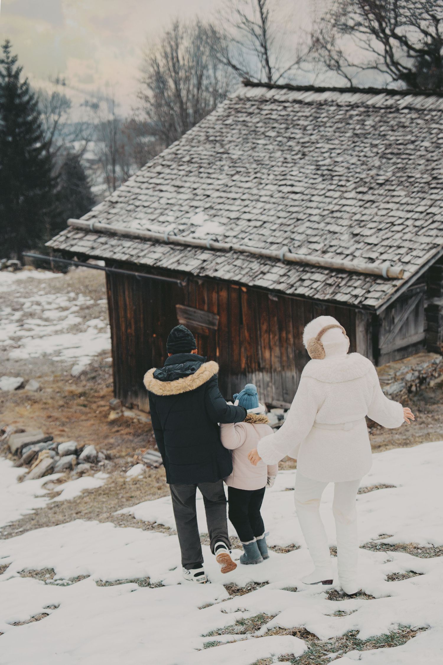 Family of three walking toward a rustic wooden chalet in a snowy alpine village.