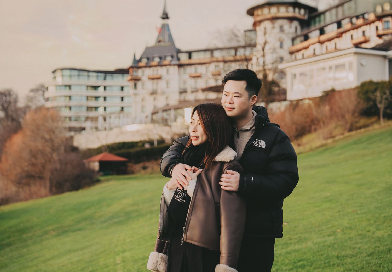 Couple embracing on green lawn with historic castle hotel in background during autumn.