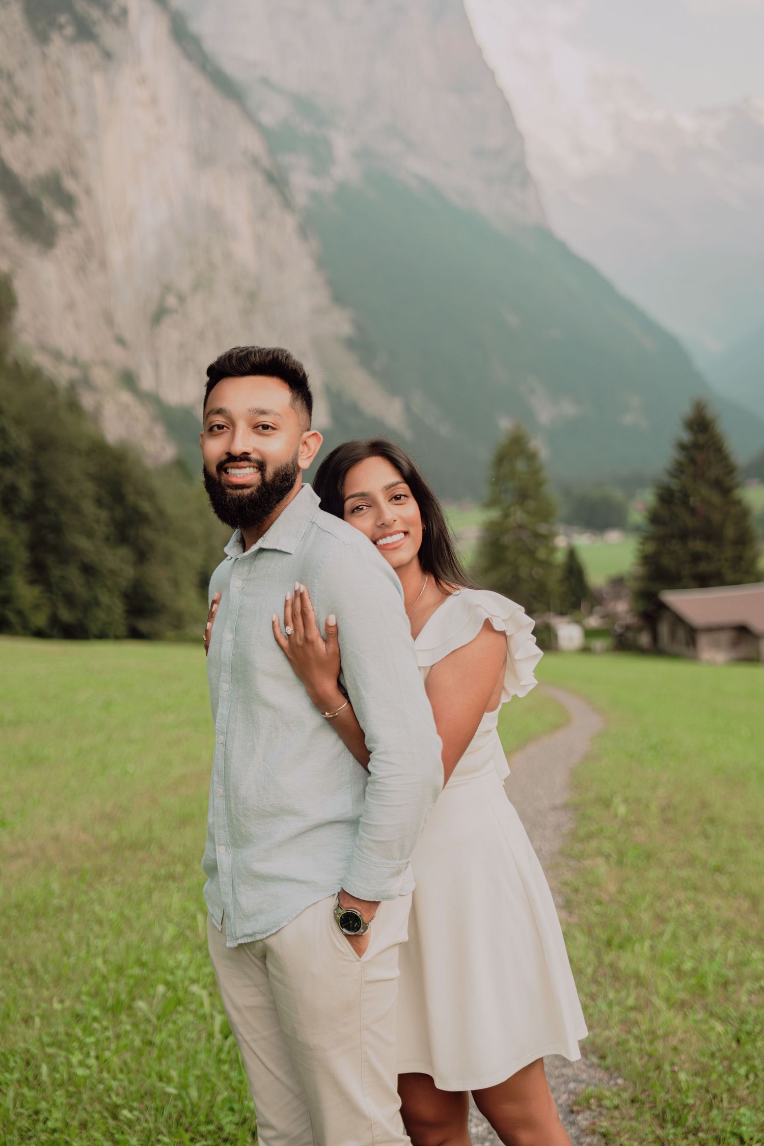 Happy engaged couple posing in a lush green alpine meadow, woman in white dress hugging man from behind.