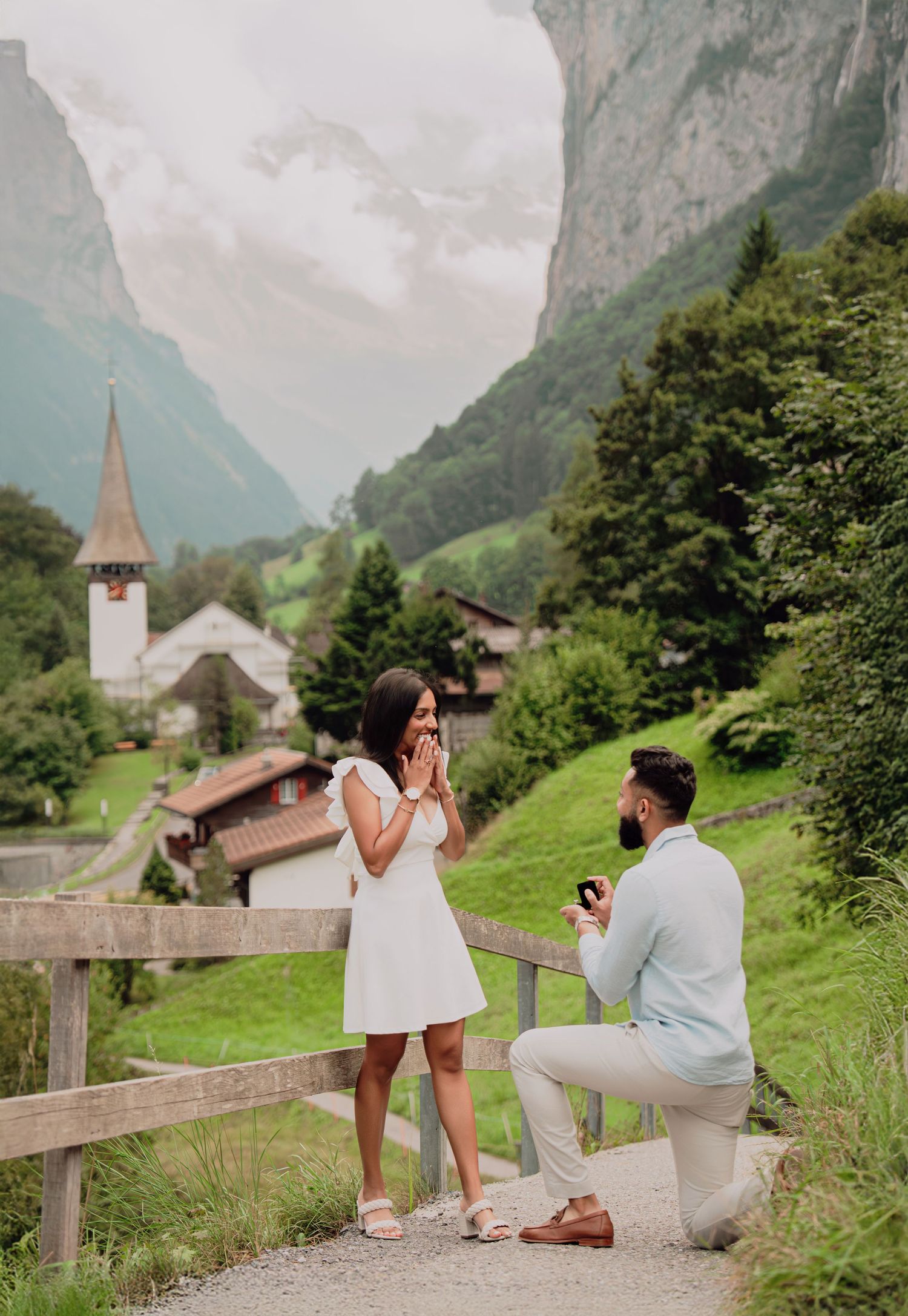 A man kneels to propose to an emotional woman on a scenic Swiss mountain path near a charming village church.