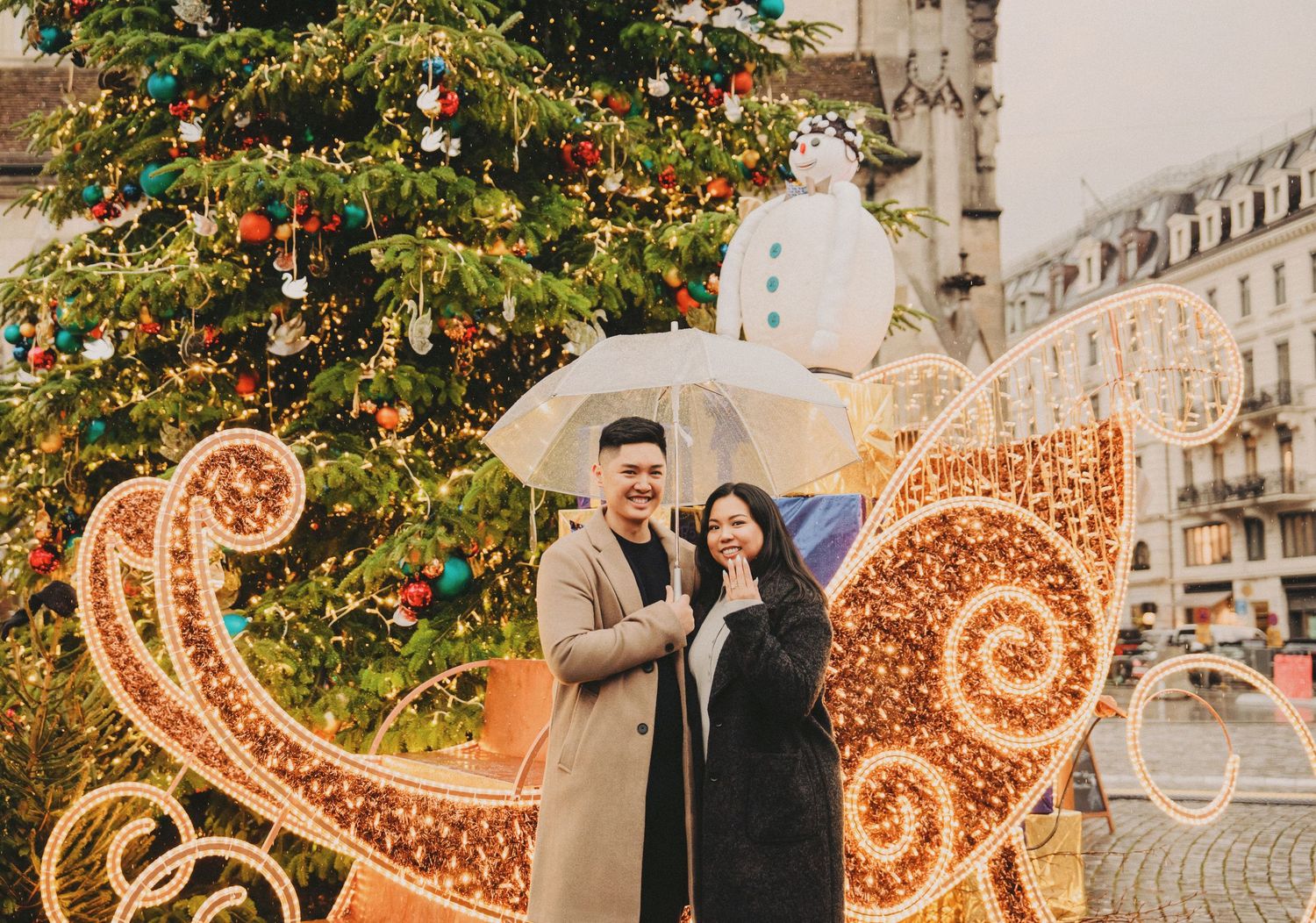 Happy couple under umbrella posing by large decorated Christmas tree.