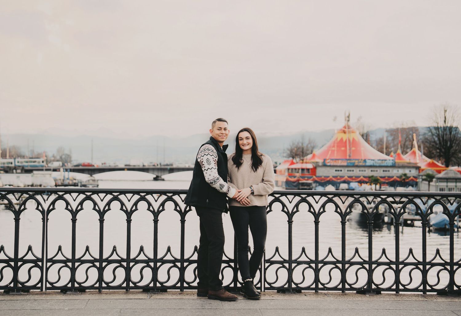 Smiling couple posing by ornate iron railings with a colorful circus tent and lake visible in Zurich.